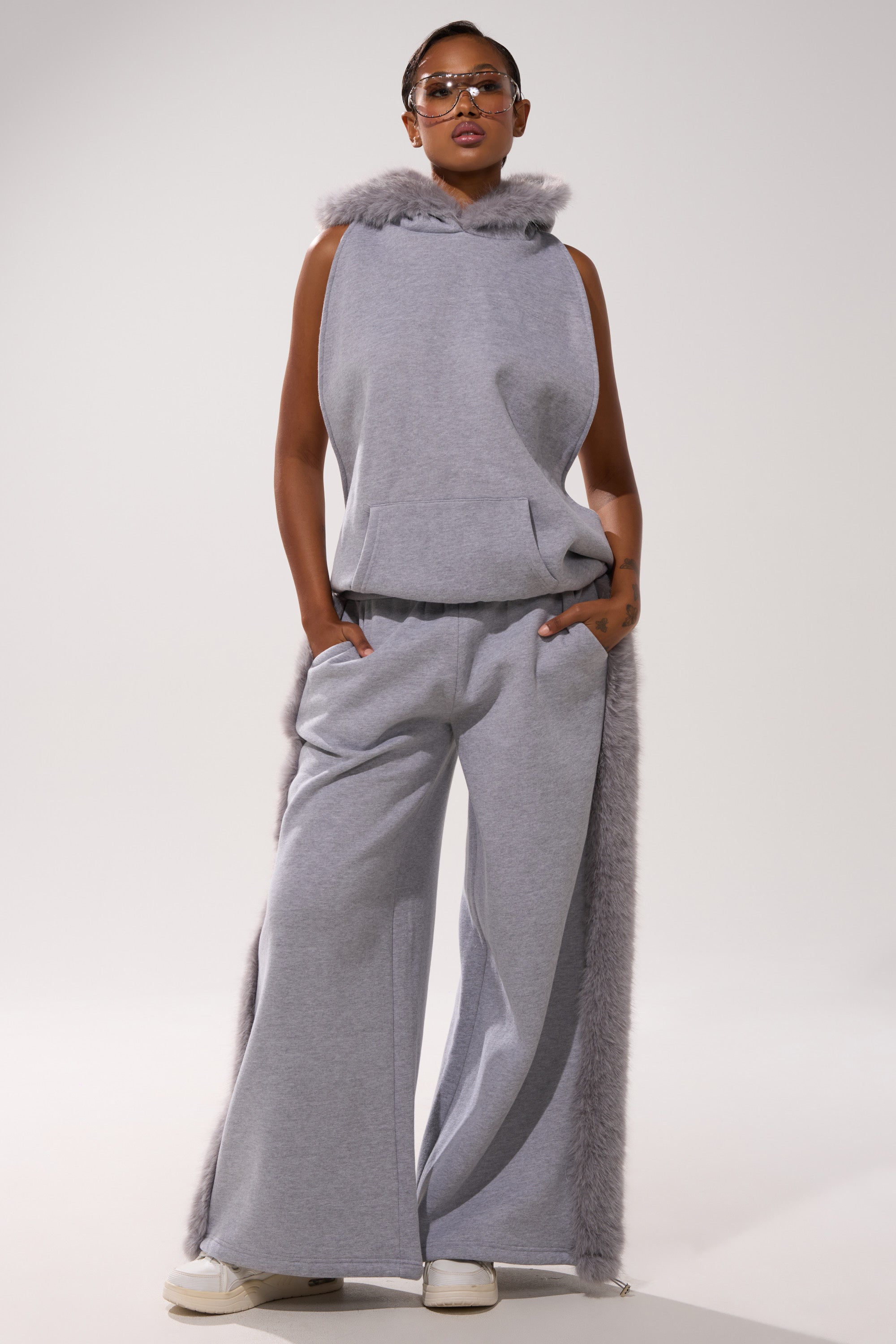 A woman stands confidently in LUXE FUR JOGGER IN GREY, styled with a sleeveless gray fur-trim hoodie, tinted sunglasses, and white sneakers, hands in her pockets.