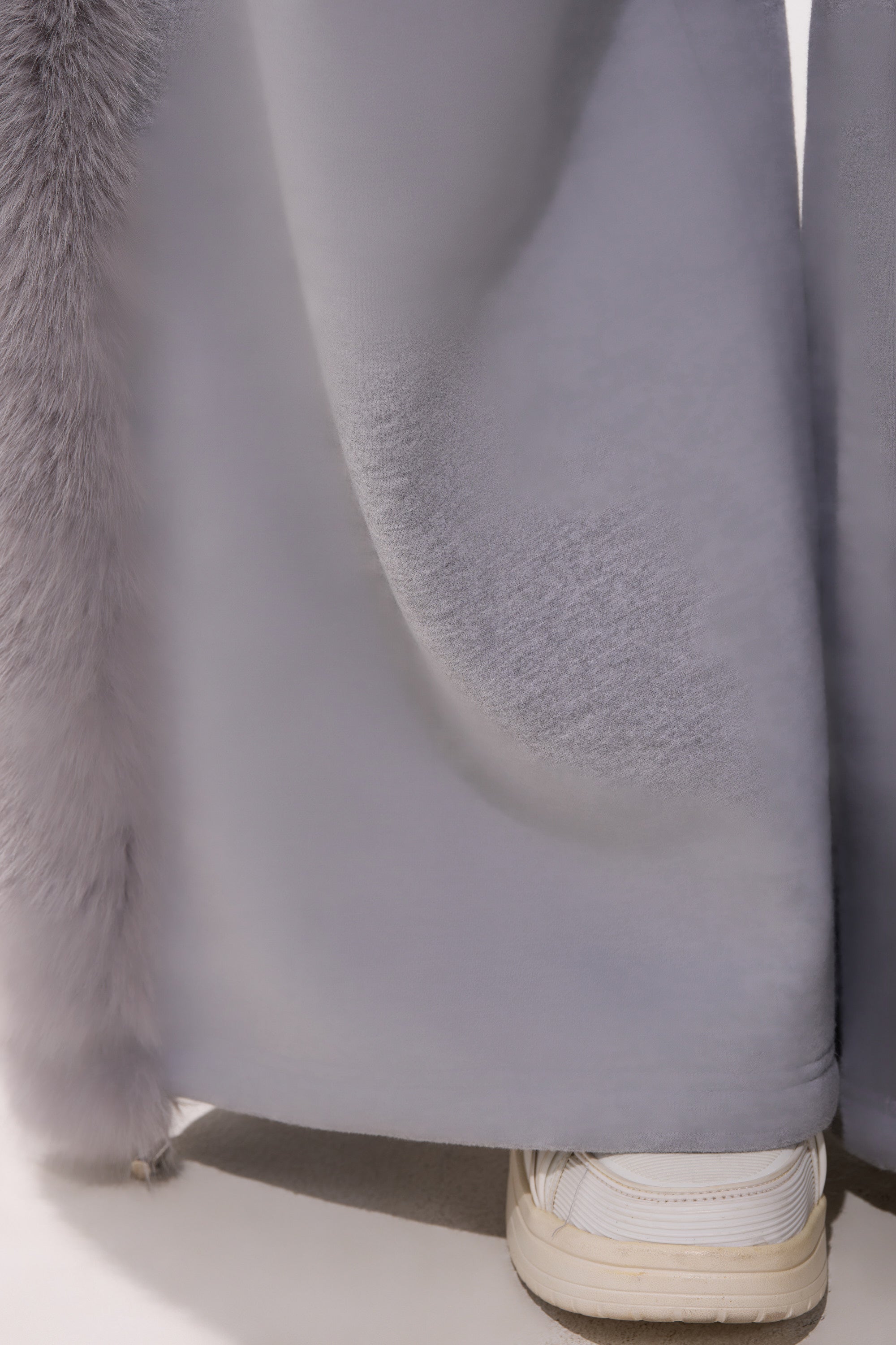 A close-up of the hem of a light gray, furry coat partially covering the LUXE FUR JOGGER IN GREY and a white sneaker on a smooth, light-colored surface.