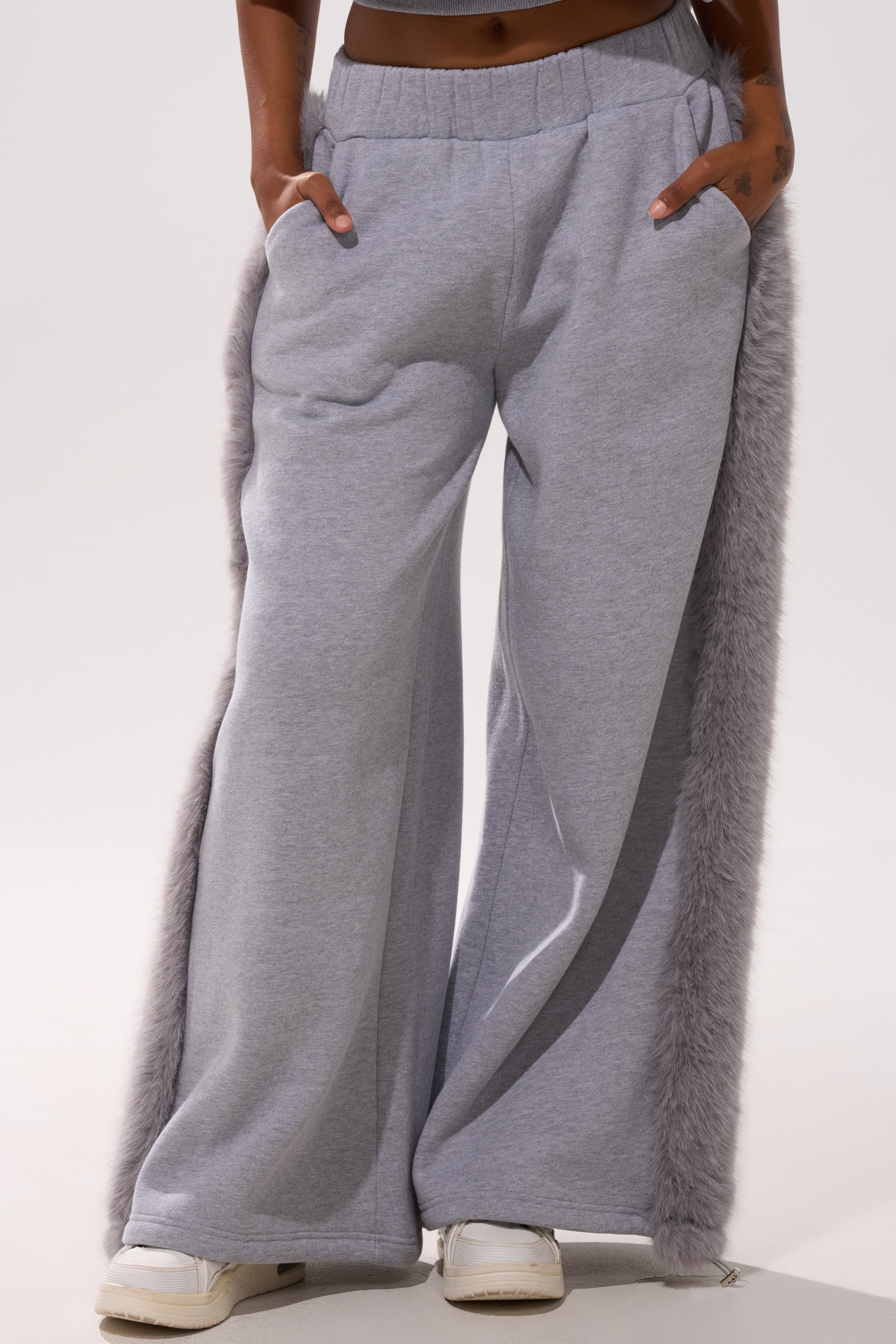 A person wearing the LUXE FUR JOGGER IN GREY, featuring wide-leg fleece and light gray faux fur trim on the sides, stands with hands in pockets and white sneakers.