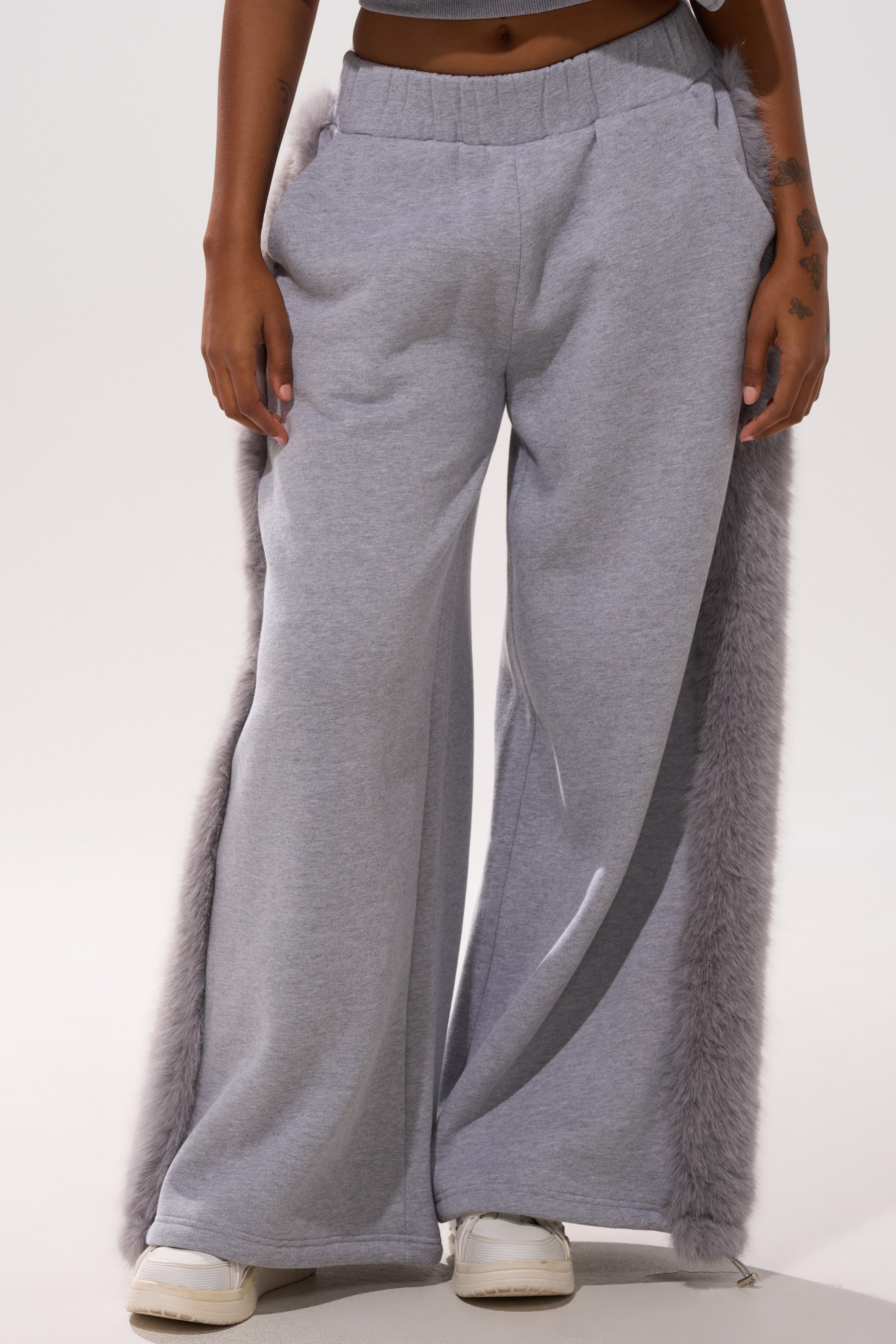 A person wears the LUXE FUR JOGGER IN GREY—light gray joggers with wide legs and plush faux fur trim on the sides—paired with white sneakers. Only their lower half is shown against a plain background.