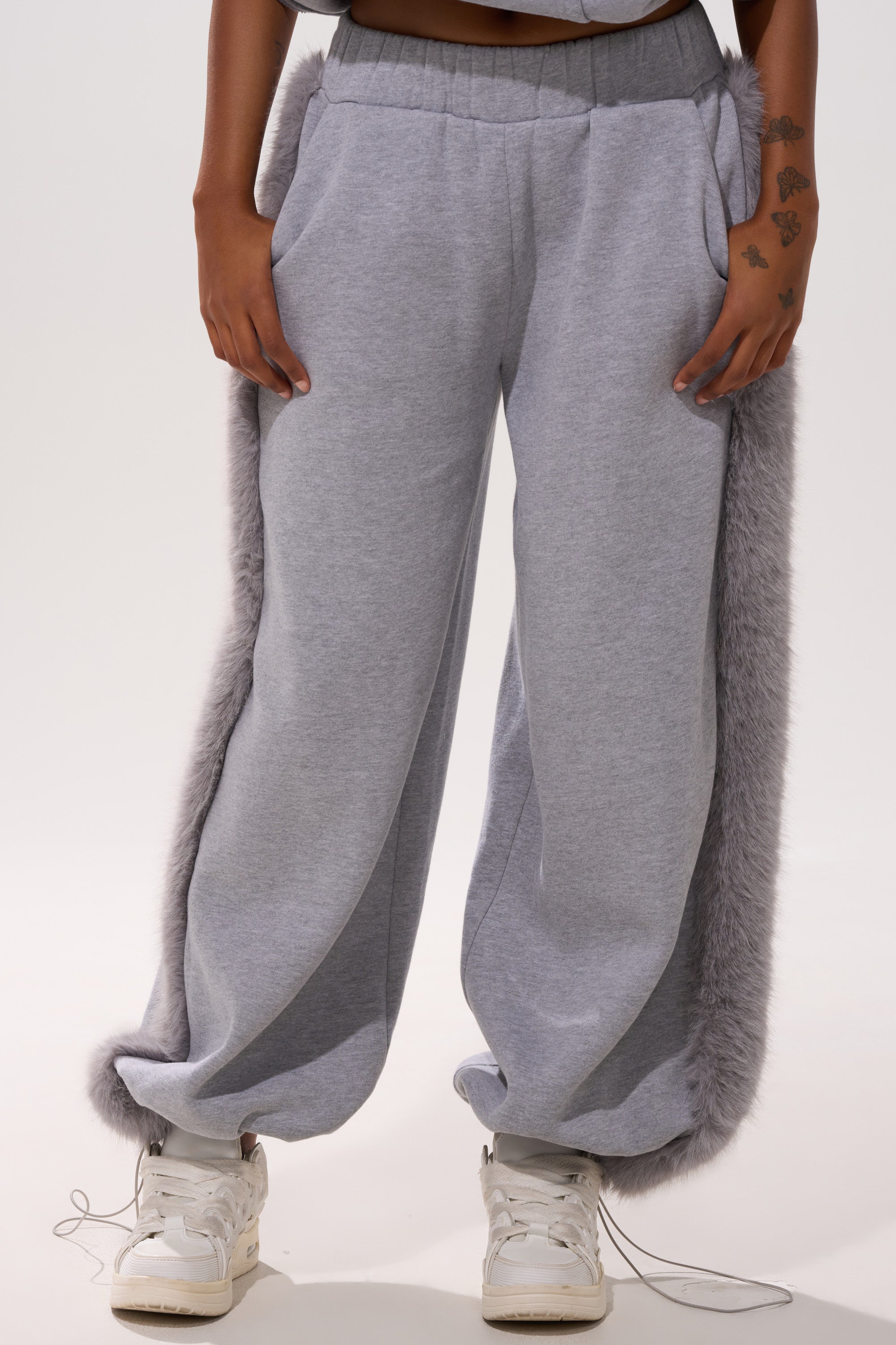 A person wears the LUXE FUR JOGGER IN GREY with fur trim on the side seams, paired with white sneakers. Their hands are in their pockets, revealing a hand tattoo, against a plain white background.