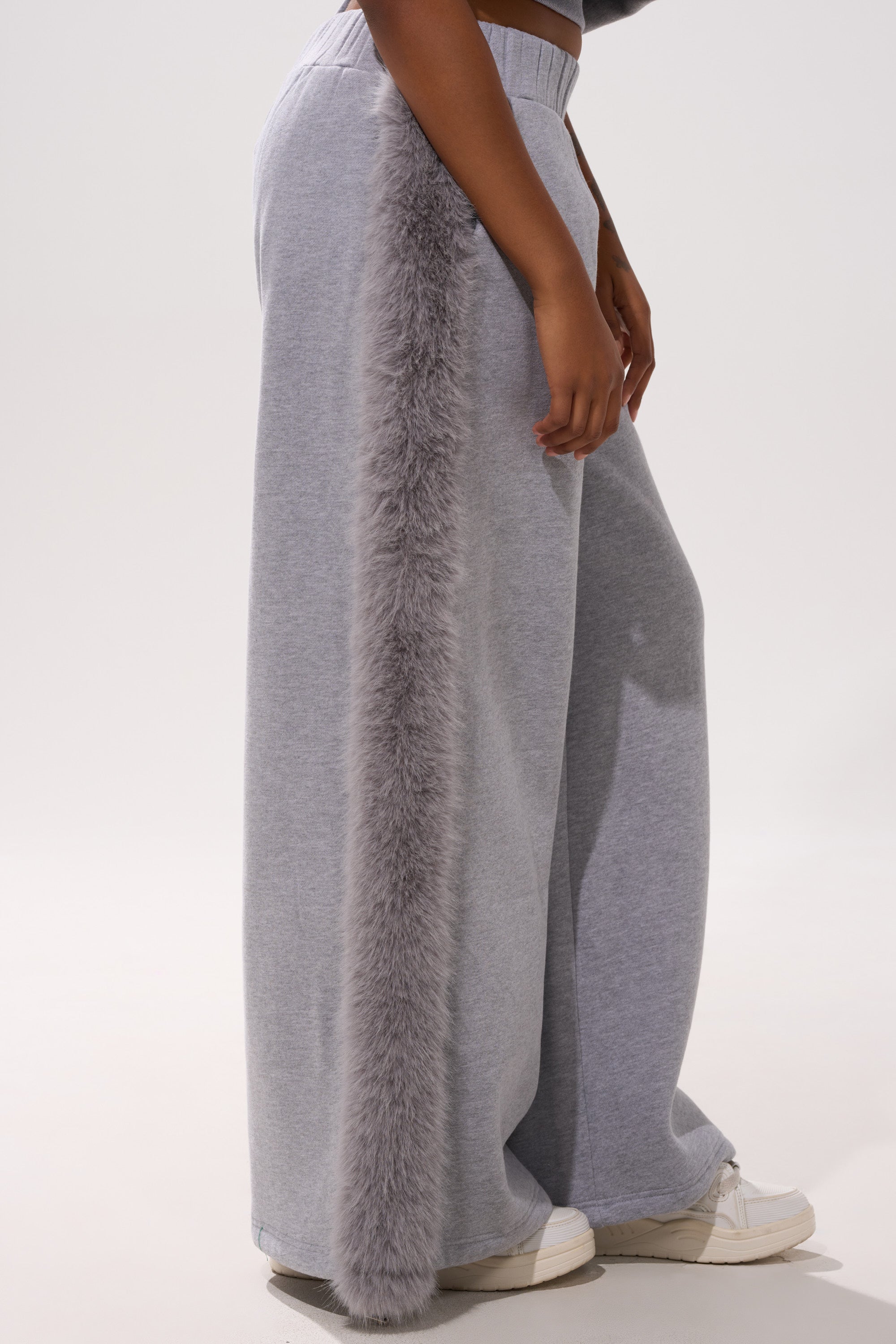 Wearing the LUXE FUR JOGGER IN GREY, cozy wide-leg sweatpants featuring a thick vertical strip of gray faux fur down the side, paired with white sneakers.