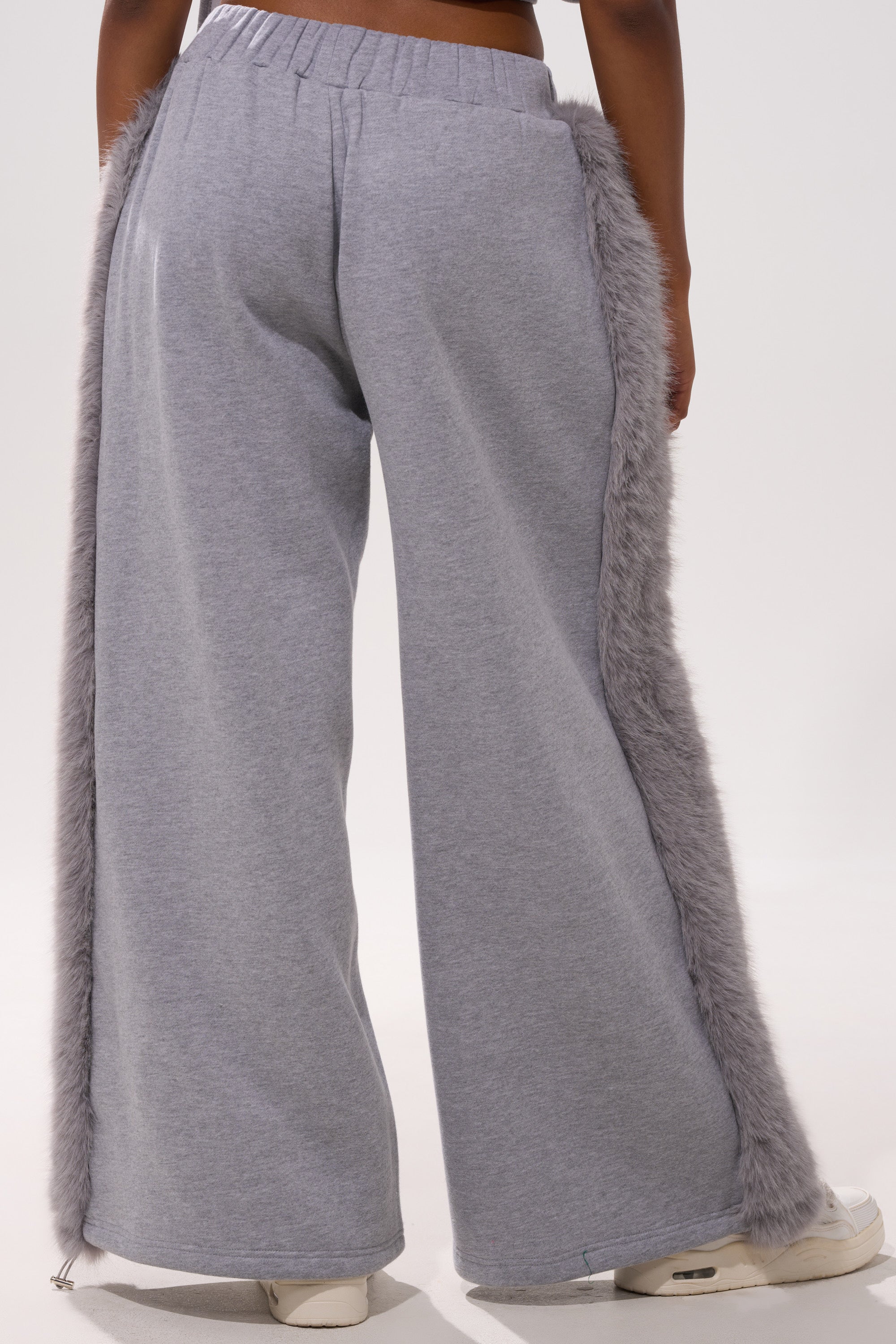 A person models the LUXE FUR JOGGER IN GREY, featuring wide legs and furry gray side trim, styled with white sneakers. The image is shot from behind, capturing only their lower torso and legs.
