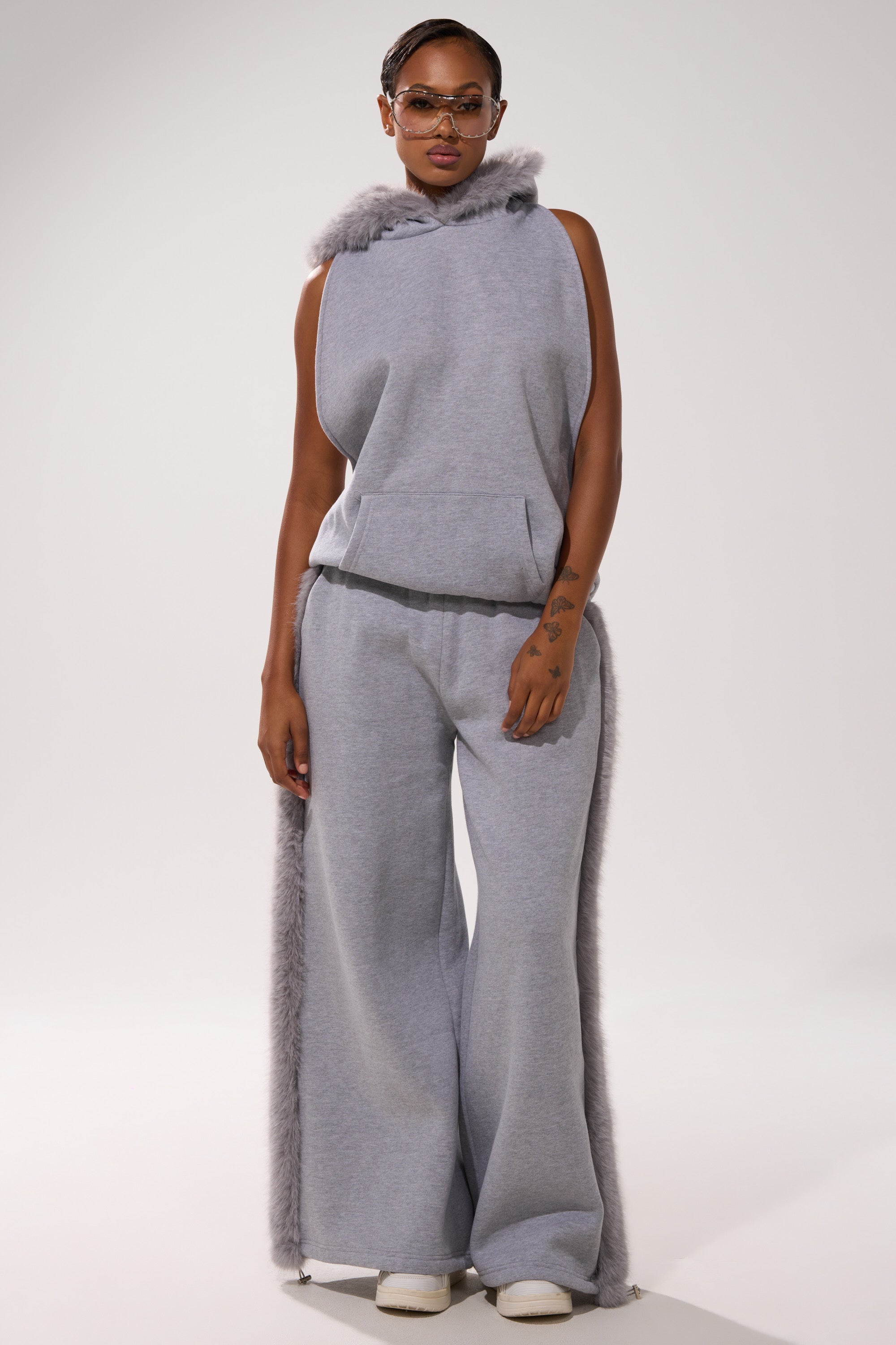 Wearing a sleeveless grey hoodie, sunglasses, white shoes, and the LUXE FUR JOGGER IN GREY with fur side trim, a person stands against a plain light background.