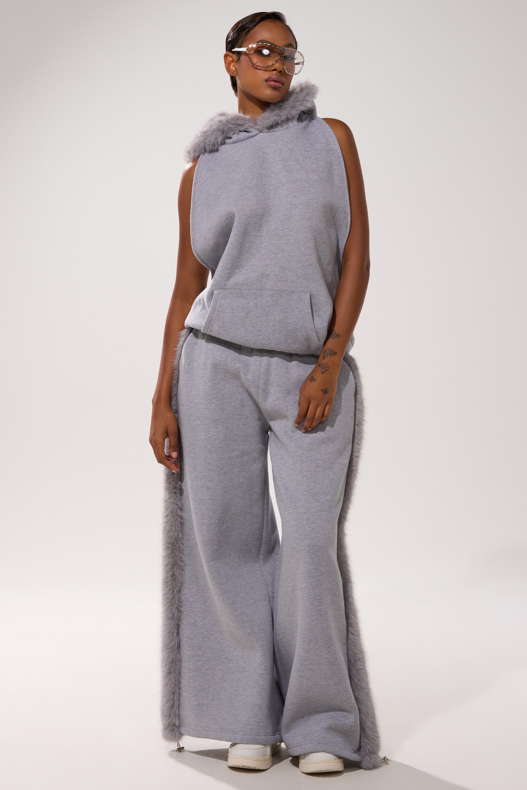 A person stands confidently in the LUXE FUR JOGGER IN GREY, a sleeveless gray hooded top with faux fur trim and matching wide leg joggers, both with chic fur accents, styled with white shoes and clear glasses.