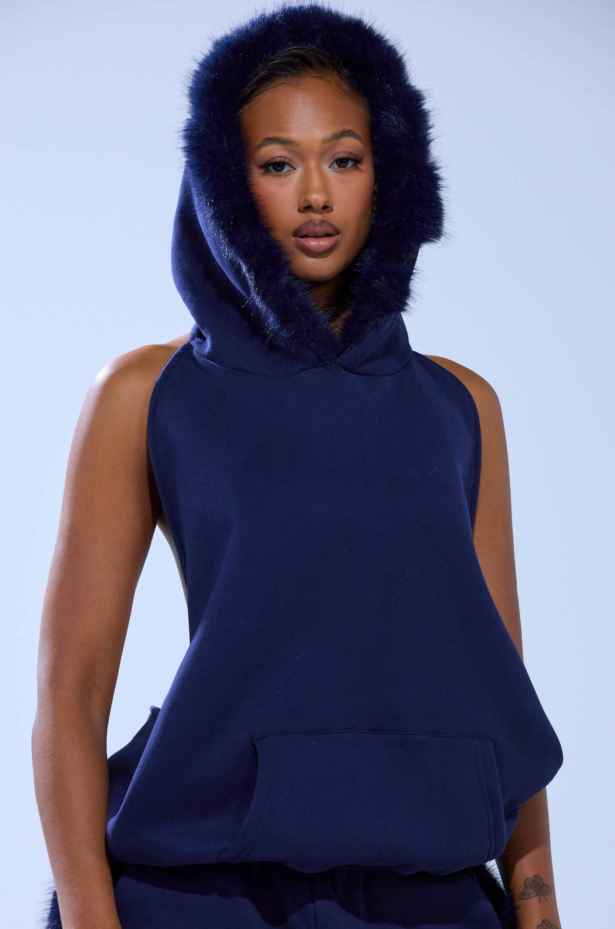 A woman with a neutral expression stands against a light blue background wearing the LUXE FUR OPEN BACK SWEATSHIRT IN NAVY featuring faux fur trim, her hands resting at her sides.