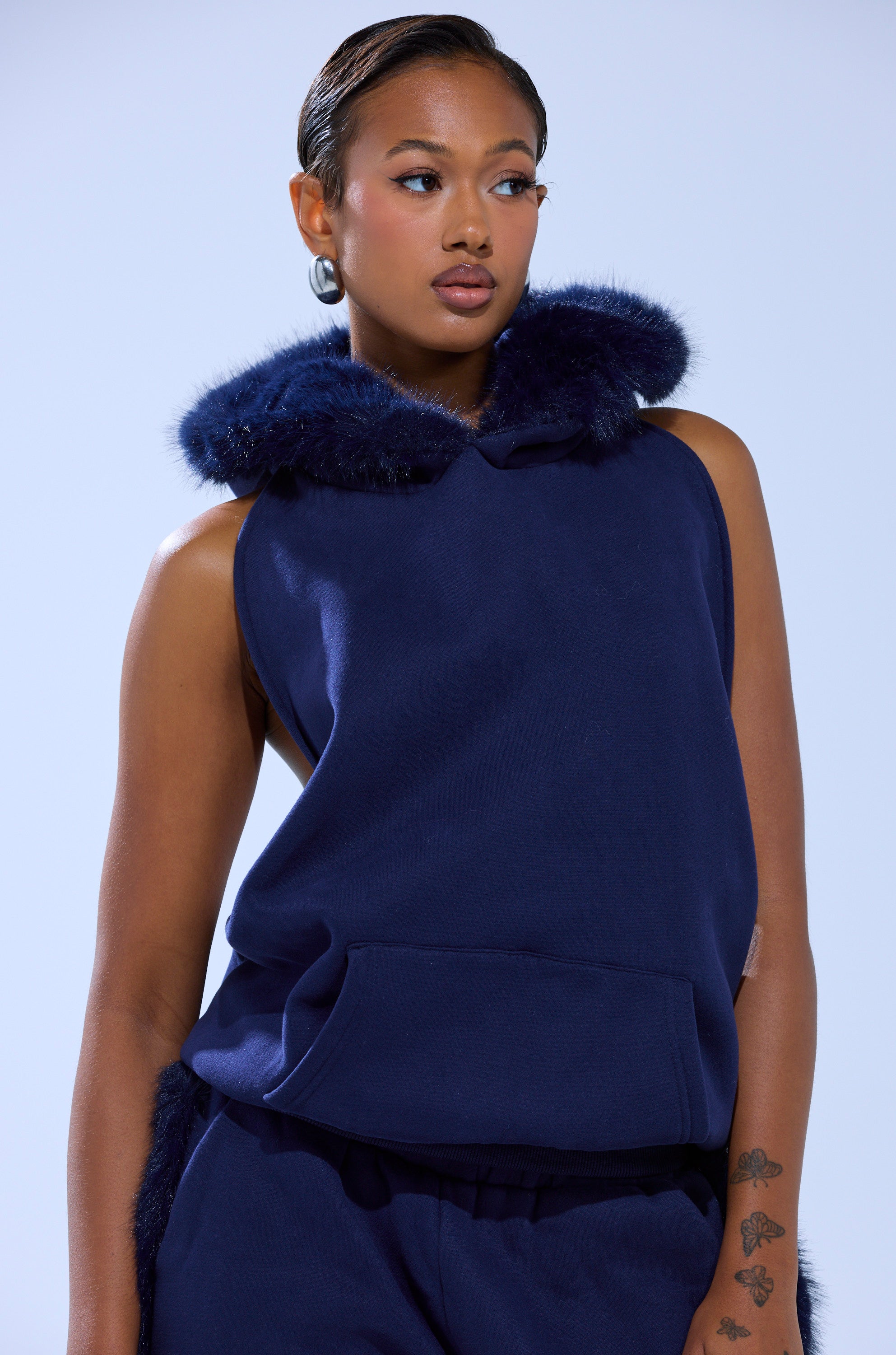 A person in a sleeveless navy LUXE FUR OPEN BACK SWEATSHIRT with faux fur hood trim and matching pants stands against a light background, looking to the side.