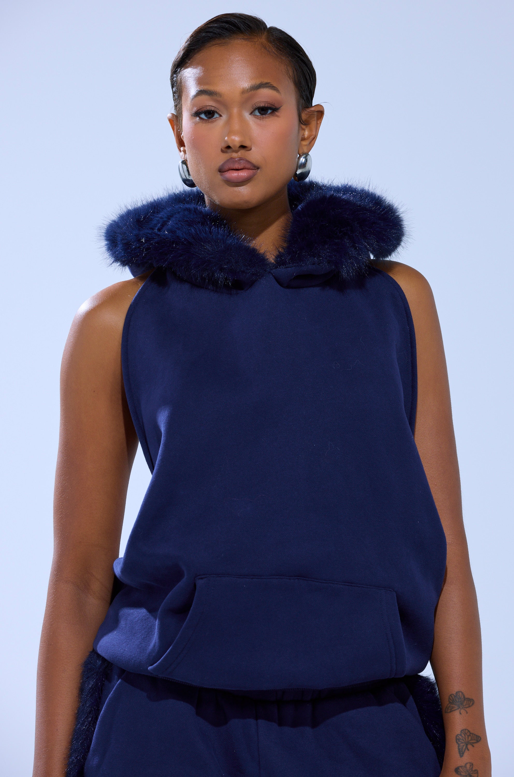 A person in the LUXE FUR OPEN BACK SWEATSHIRT IN NAVY—with faux fur-trimmed hood—styled with hoop earrings and a short, sleek hairstyle poses against a light background.