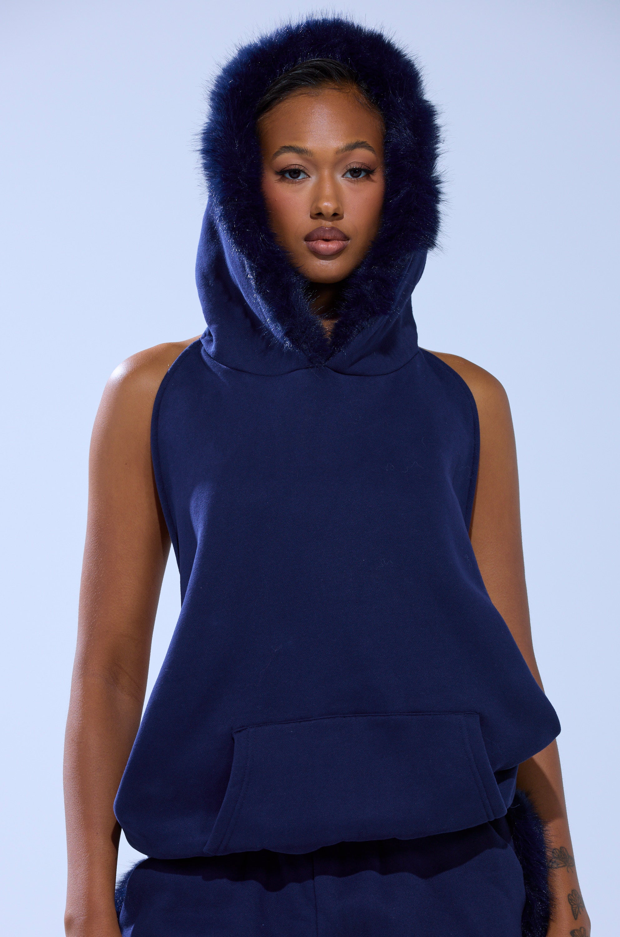A woman models the LUXE FUR OPEN BACK SWEATSHIRT IN NAVY, featuring a sleeveless design and faux fur-trimmed hood. She stands against a light background, gazing at the camera with a neutral expression and a tattoo on her left arm.
