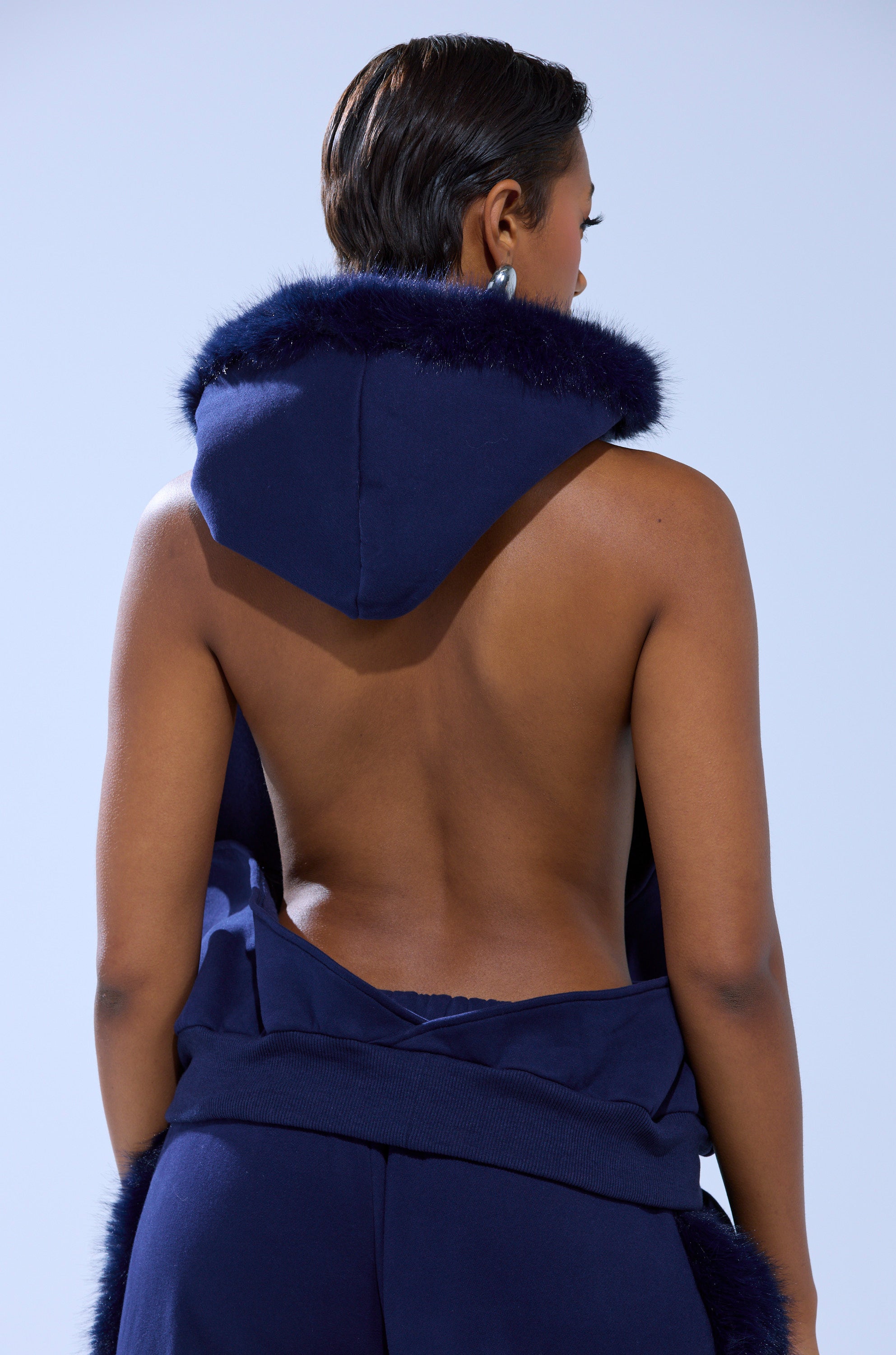Seen from behind, a person with short hair wears navy blue pants and the LUXE FUR OPEN BACK SWEATSHIRT IN NAVY, featuring faux fur trim and an open back that reveals bare shoulders, set against a light blue background.