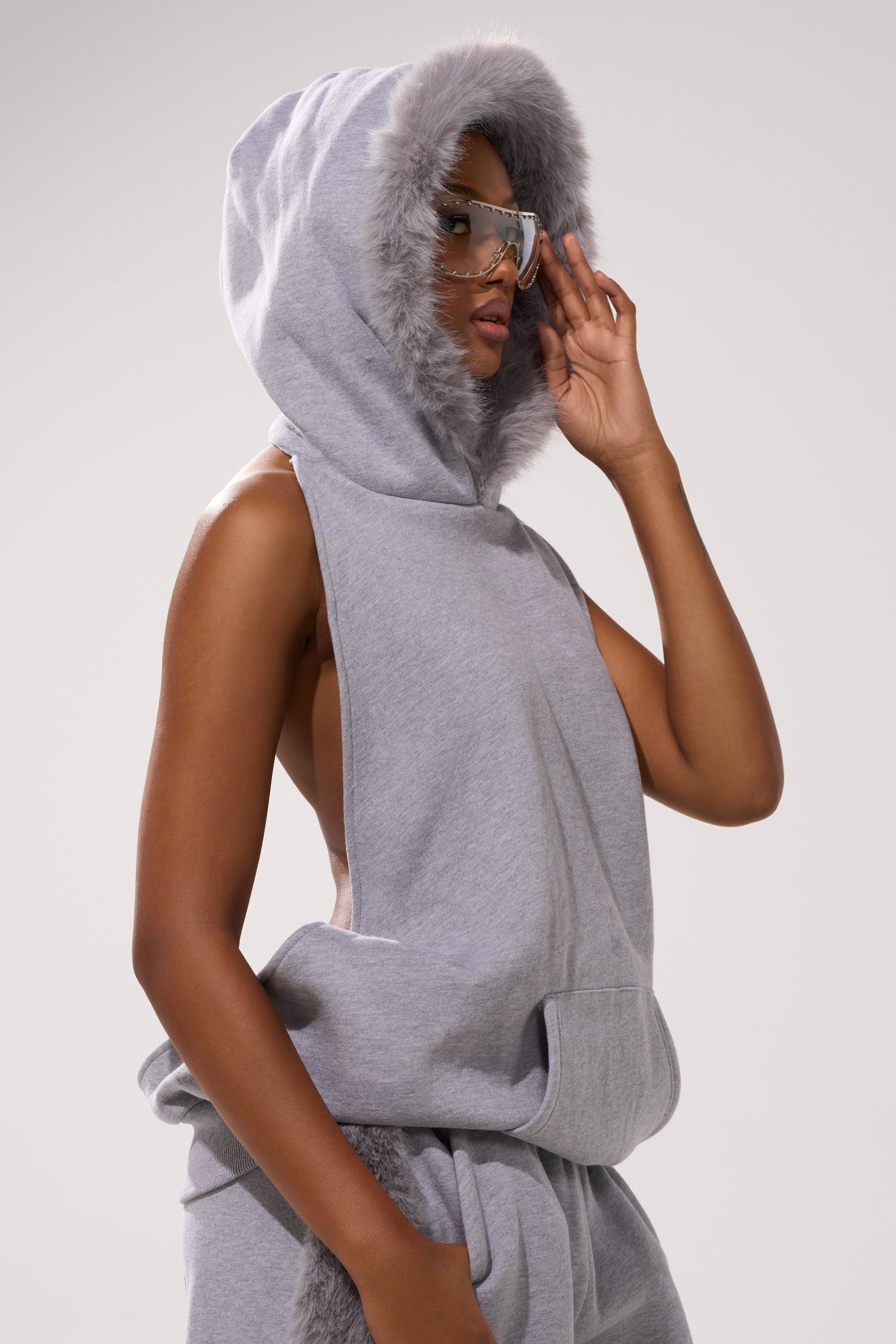 Wearing the LUXE FUR OPEN-BACK SWEATSHIRT IN GREY with a hood trimmed in faux fur and matching sweatpants, a person poses against a plain light background accessorized with large reflective sunglasses.