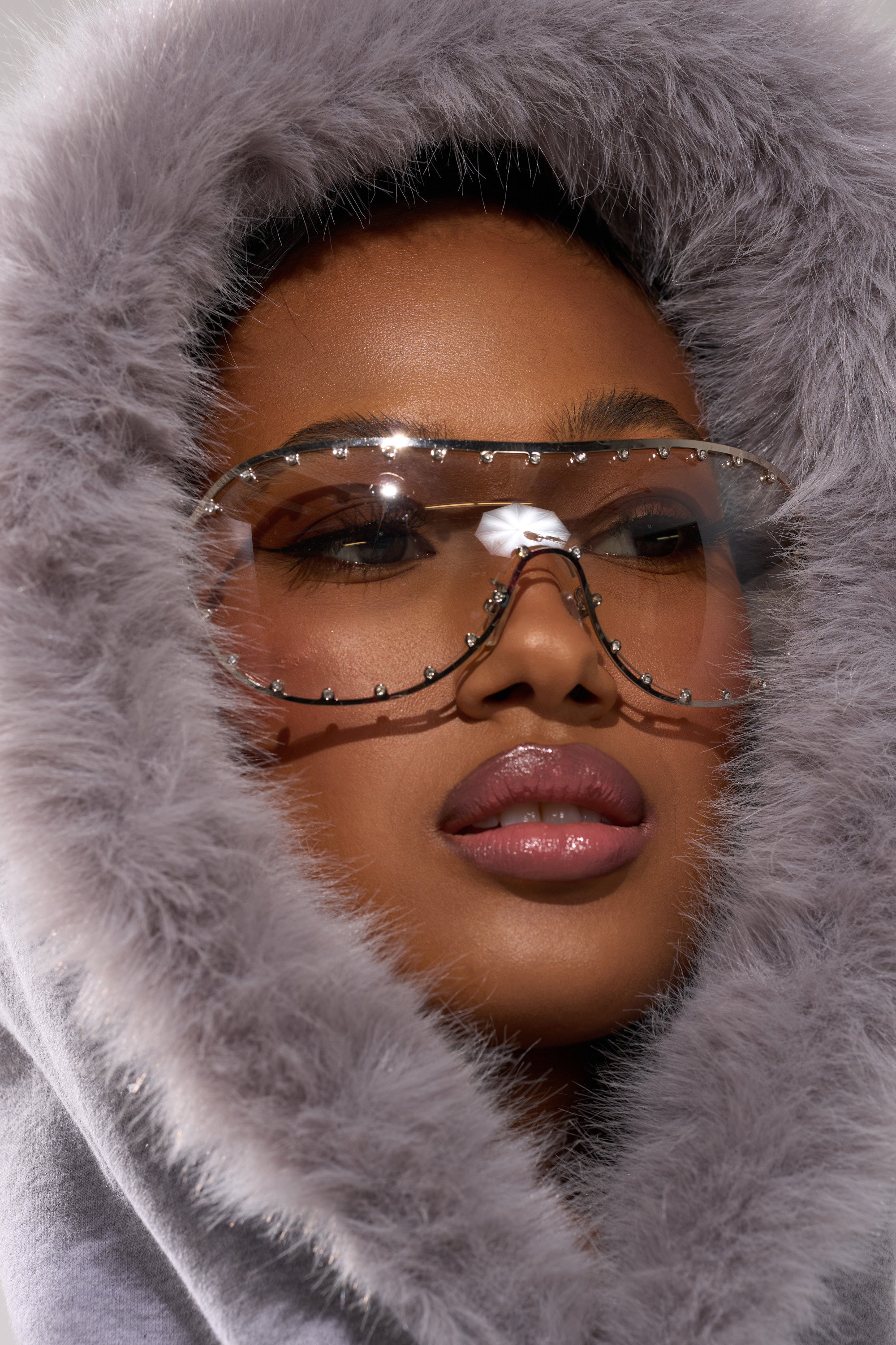 A close-up of a woman in the LUXE FUR OPEN-BACK SWEATSHIRT IN GREY, wearing large, clear, rhinestone-studded sunglasses and highlighting her smooth skin and glossy lips.