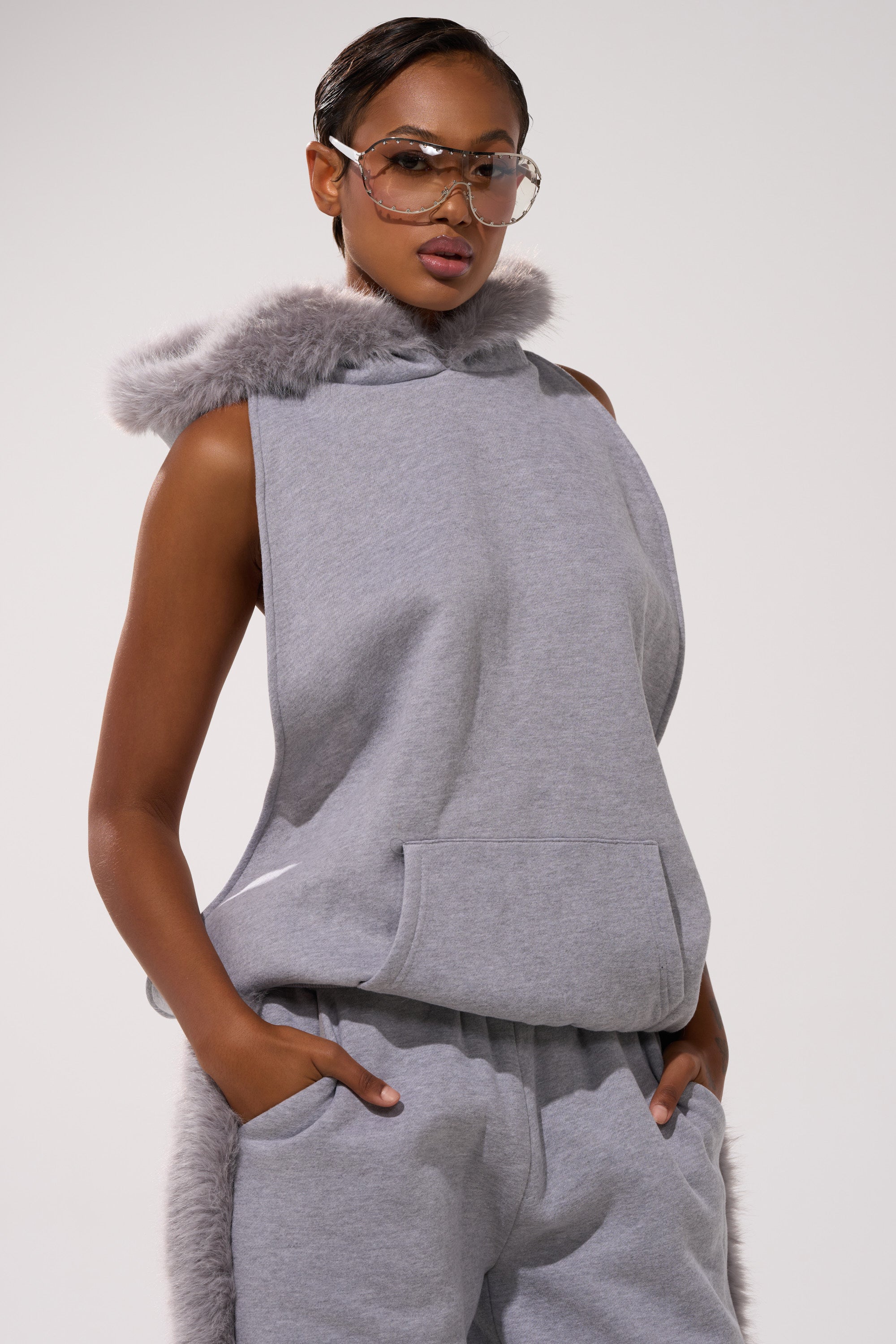 A person wearing the LUXE FUR OPEN-BACK SWEATSHIRT IN GREY, featuring a luxe fur-trimmed hood, pairs it with matching sweatpants. They have short, sleek hair and wear oversized glasses, standing with hands in pockets.