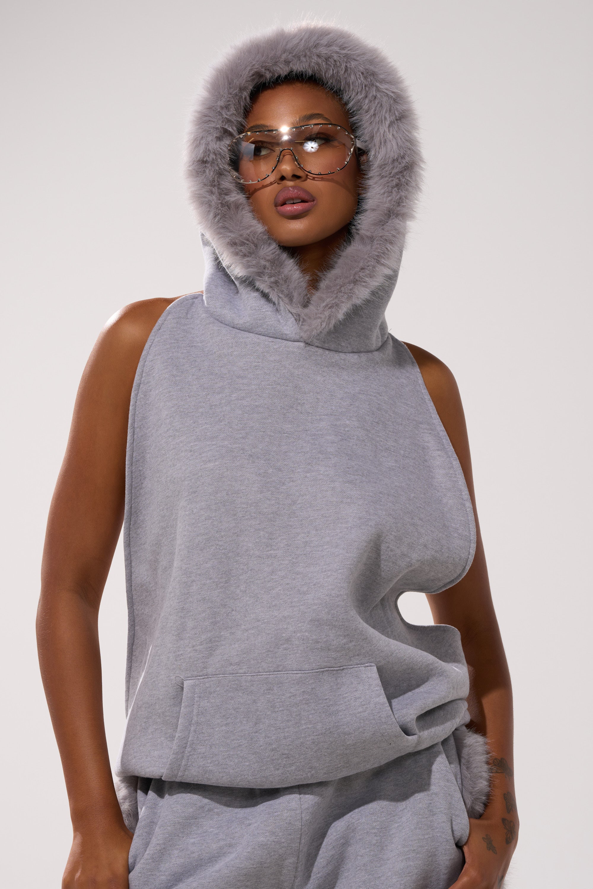 Wearing the LUXE FUR OPEN-BACK SWEATSHIRT IN GREY, matching pants, and large clear glasses, a person stands confidently against a plain background.