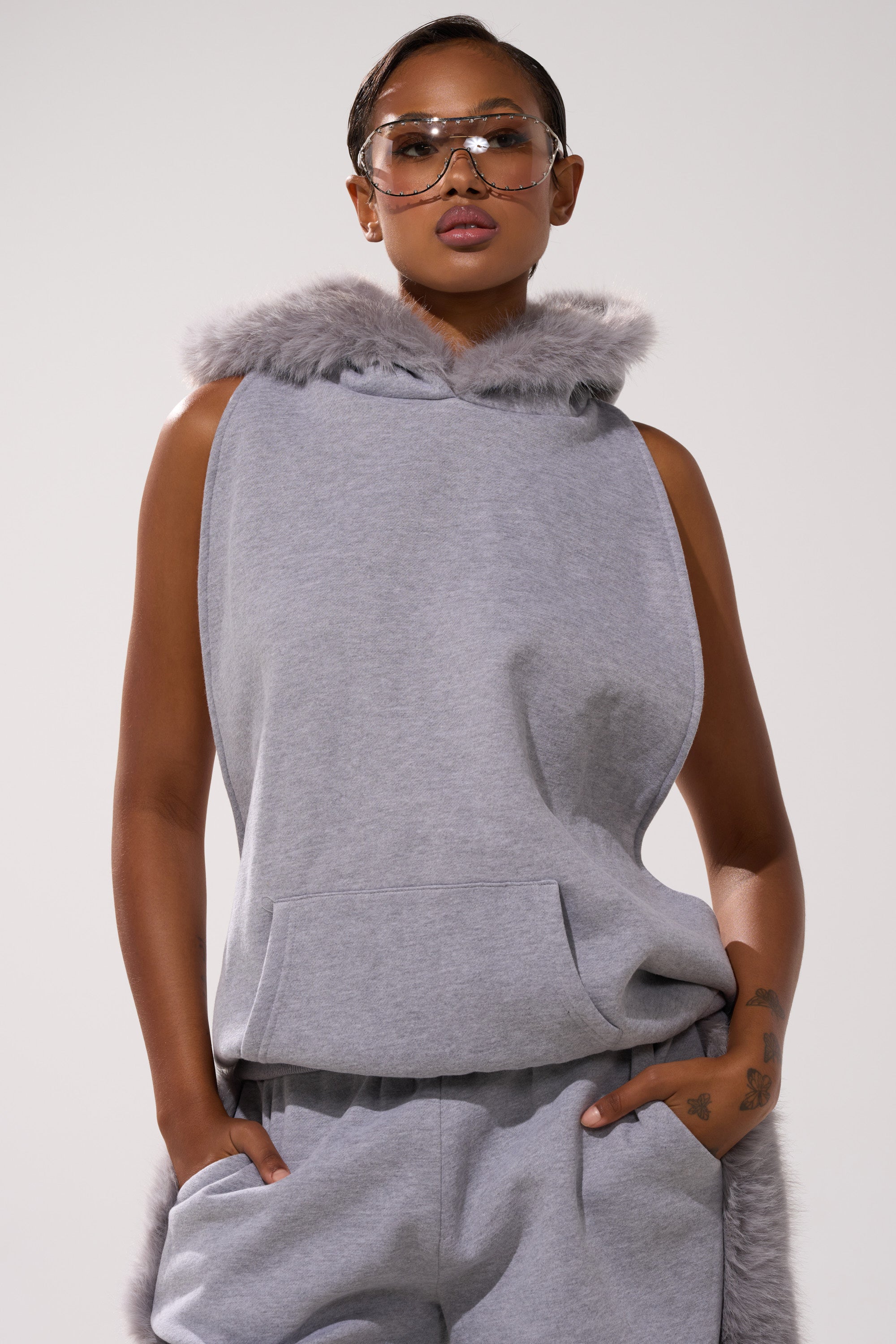 Clad in the LUXE FUR OPEN-BACK SWEATSHIRT IN GREY and matching sweatpants, a model strikes a confident pose with one hand in a pocket, finished off with bold, clear-framed sunglasses.