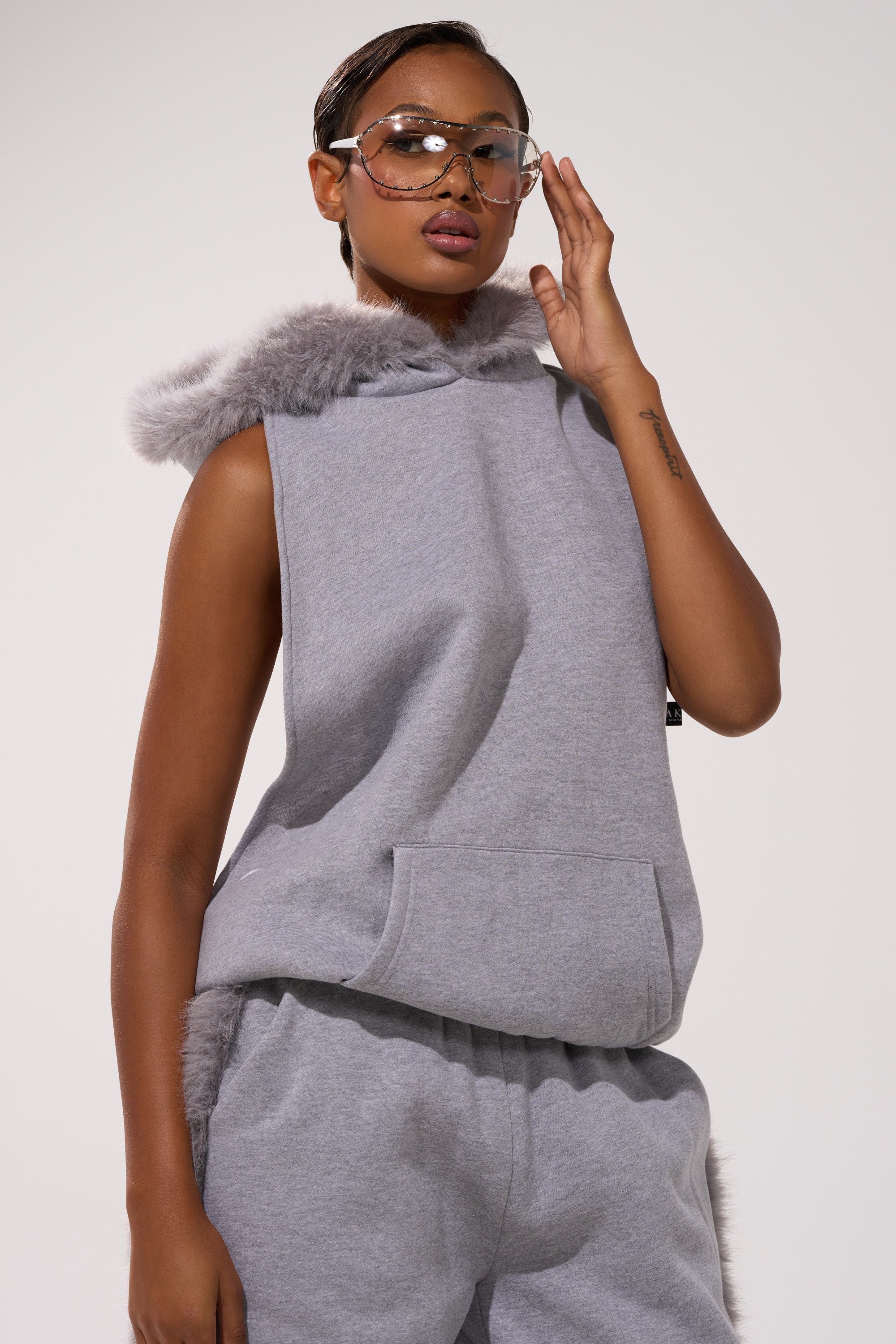 A person poses confidently against a plain background wearing the LUXE FUR OPEN-BACK SWEATSHIRT IN GREY and clear glasses, paired with matching sweatpants.