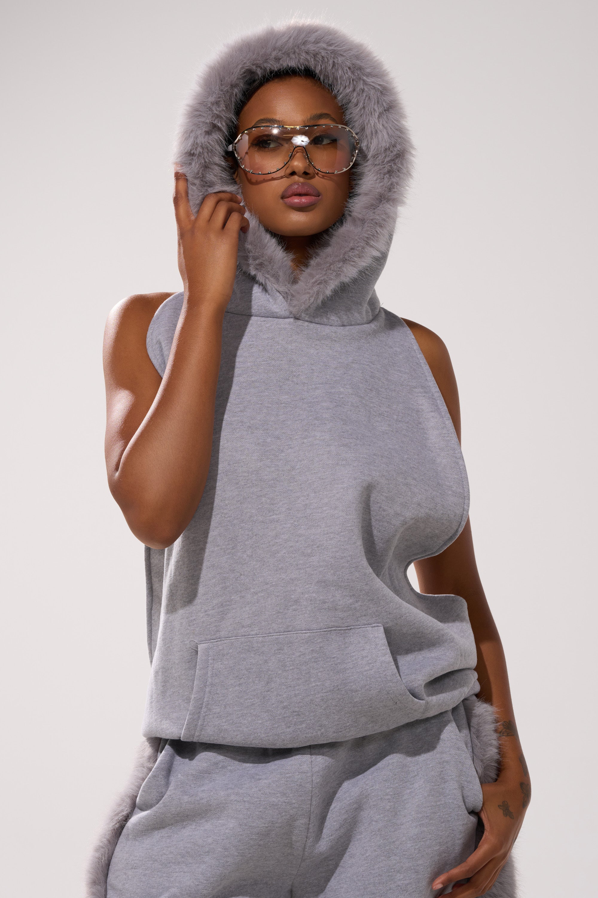 A person confidently poses in the LUXE FUR OPEN-BACK SWEATSHIRT IN GREY with matching sweatpants, accessorized with oversized sunglasses and touching the sweatshirt's hood against a neutral background.