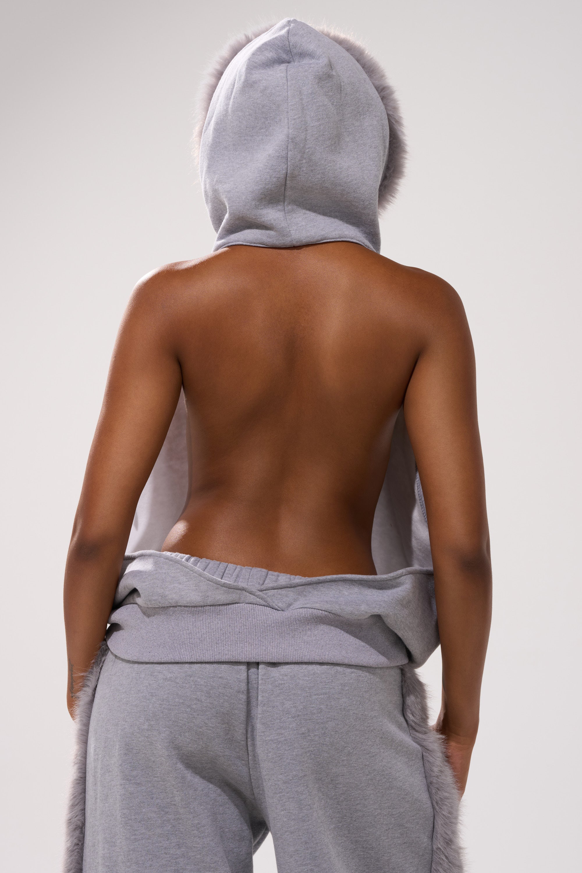A person faces away from the camera in the LUXE FUR OPEN-BACK SWEATSHIRT IN GREY, hood up, with a bare back visible. They wear matching jogger pants against a plain light background.
