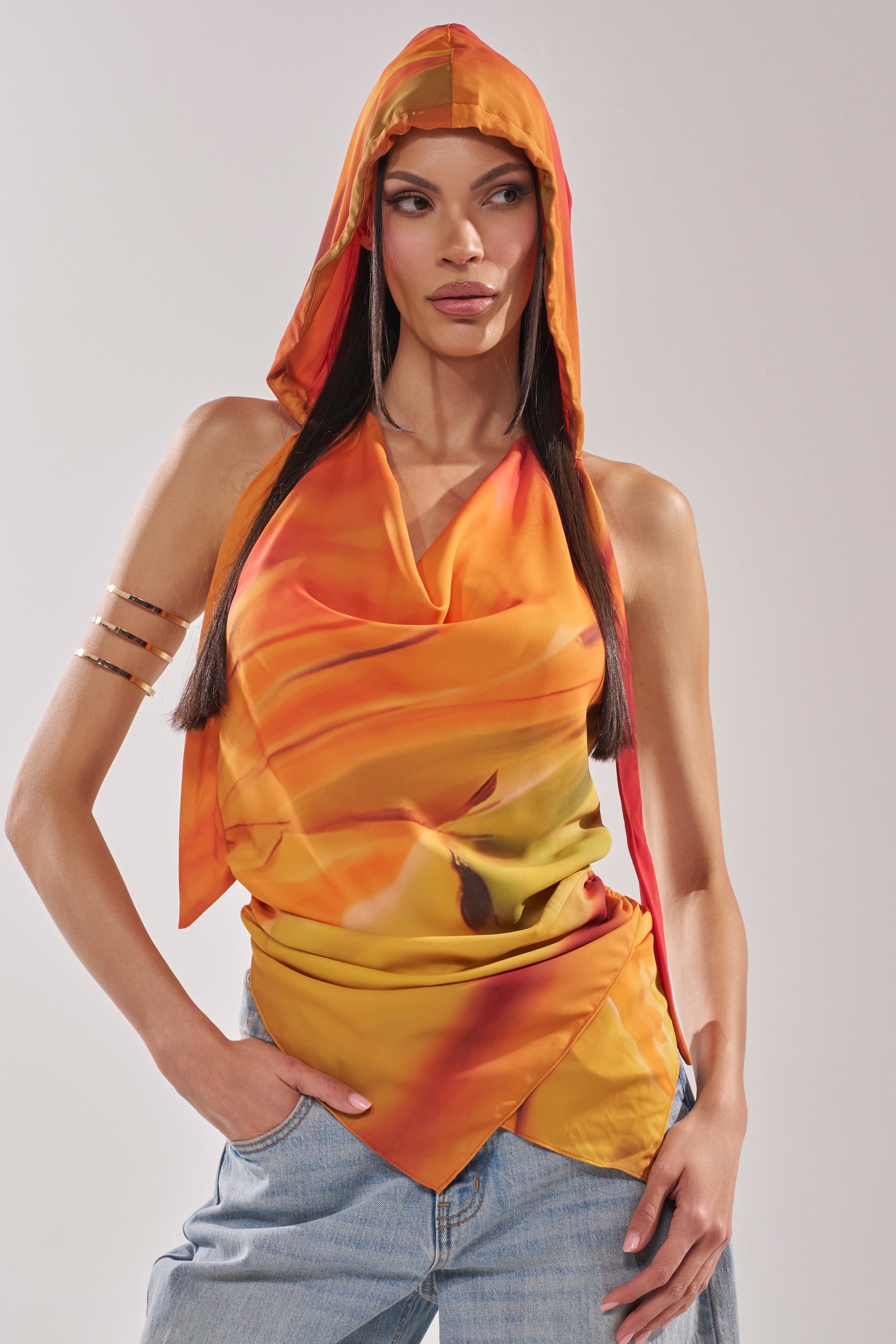 A woman poses against a light gray background, wearing the COASTAL HOODED HALTER TOP in vibrant orange and yellow satin with a draped neckline, paired with light blue jeans and a gold arm cuff, one hand in her pocket. 0