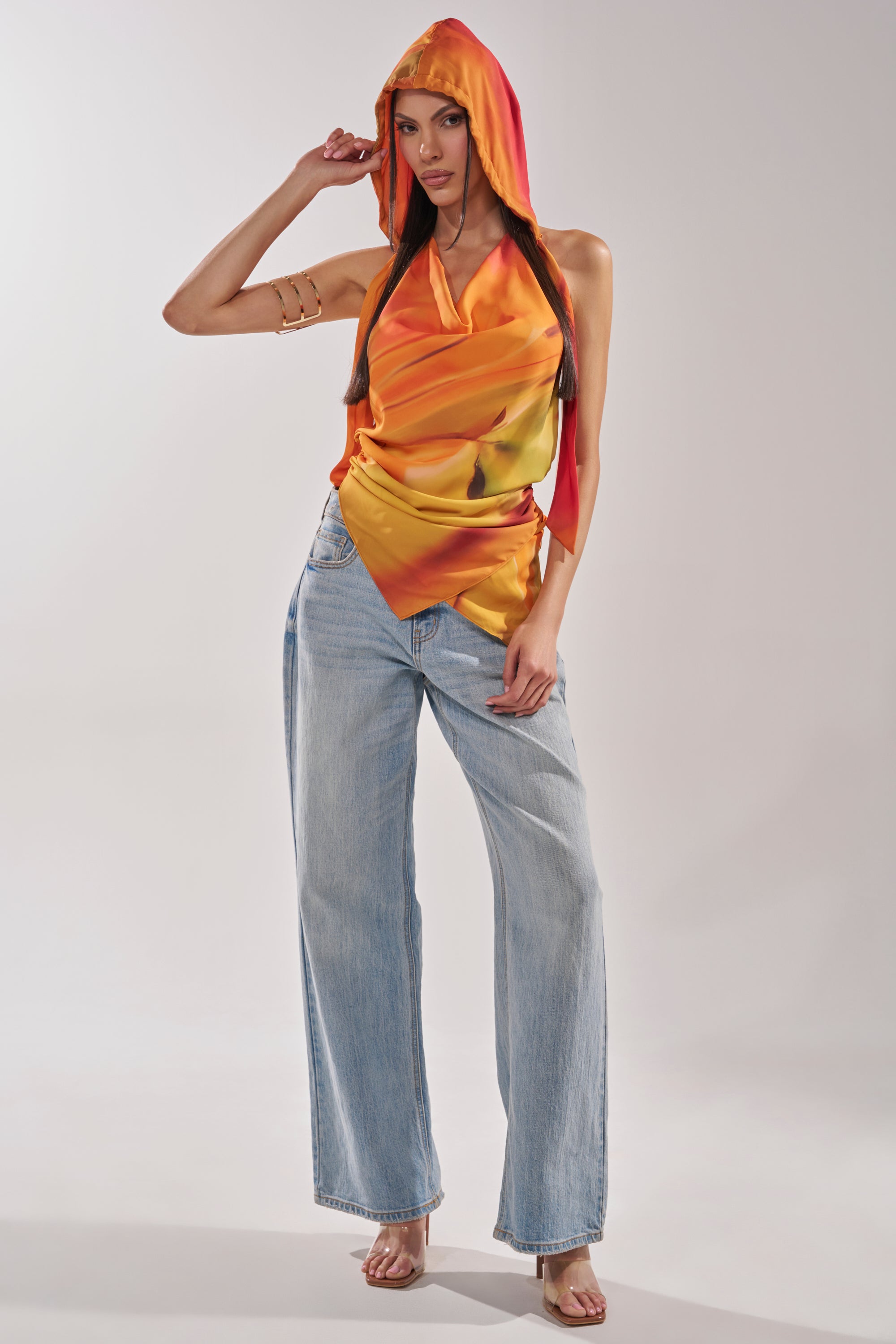 A woman poses in the COASTAL HOODED HALTER TOP, a sleeveless orange-yellow satin piece with a draped front, styled with light blue wide-leg jeans and clear heeled sandals against a plain backdrop. 9