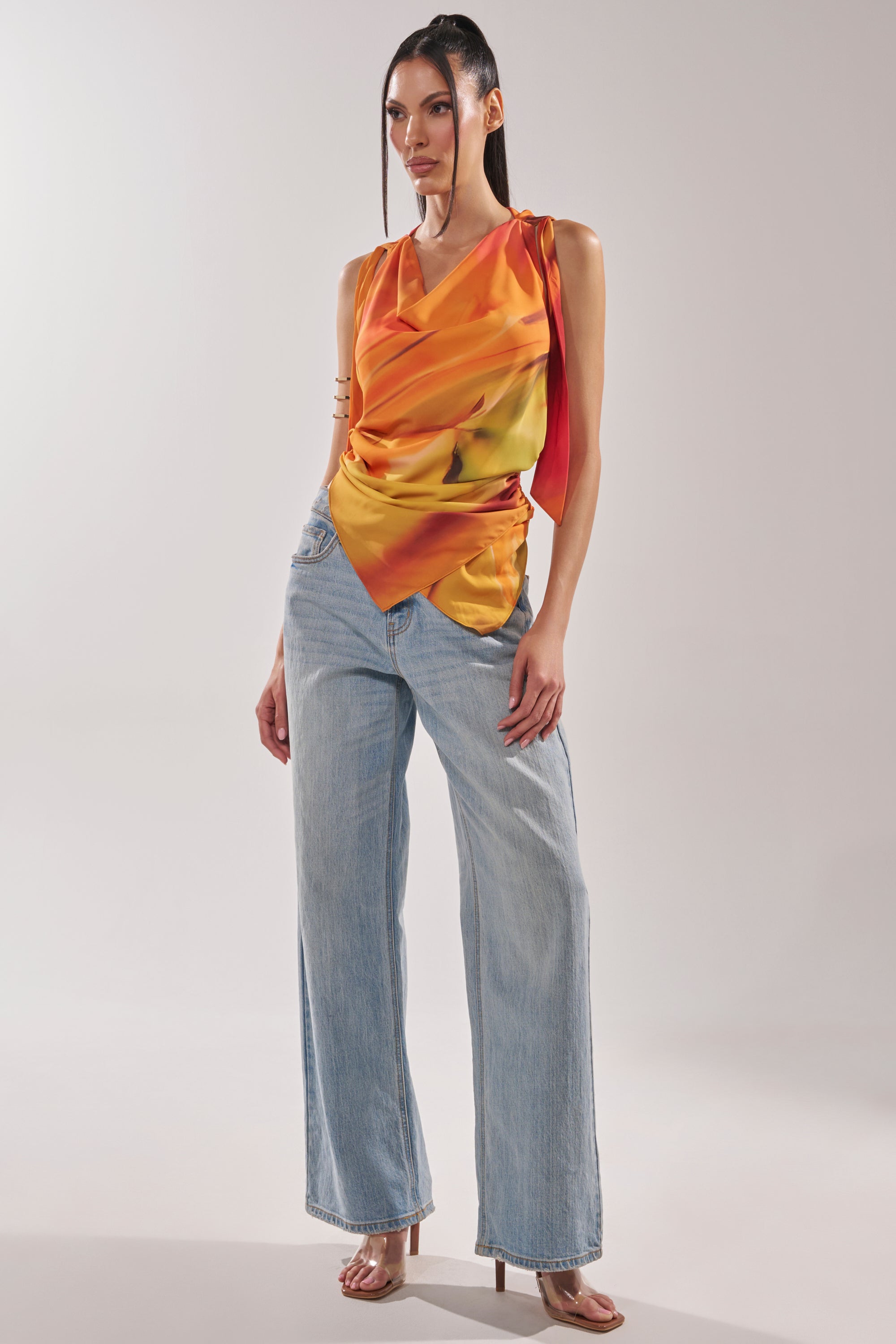 A woman poses confidently in a studio wearing the COASTAL HOODED HALTER TOP, styled with light blue wide-leg jeans and heeled sandals. Her straight hair is parted to the side as she stands with one hand on her hip. 10