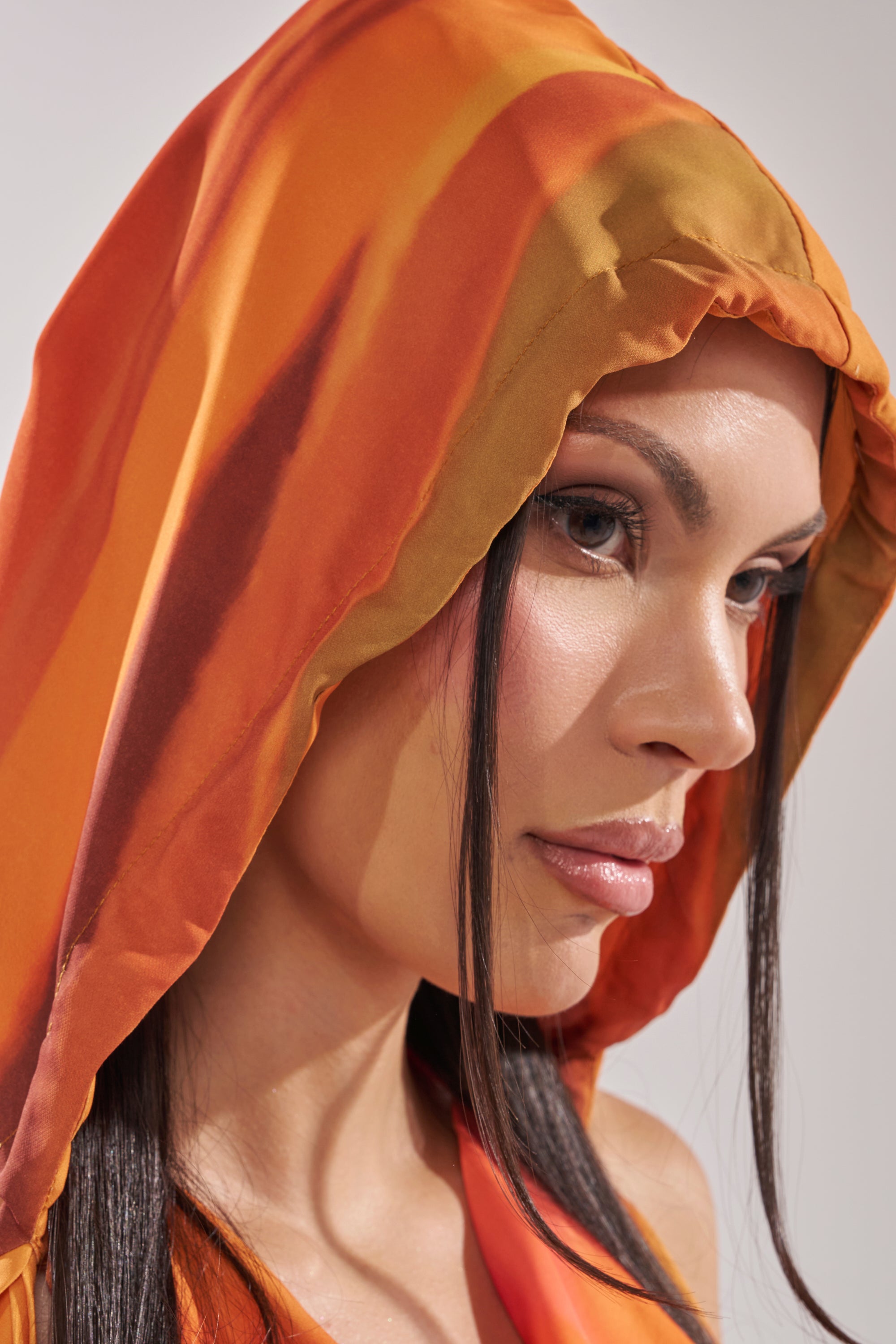 A woman with long dark hair and natural makeup looks thoughtfully to the side, wearing the COASTAL HOODED HALTER TOP in orange against a neutral background. 11