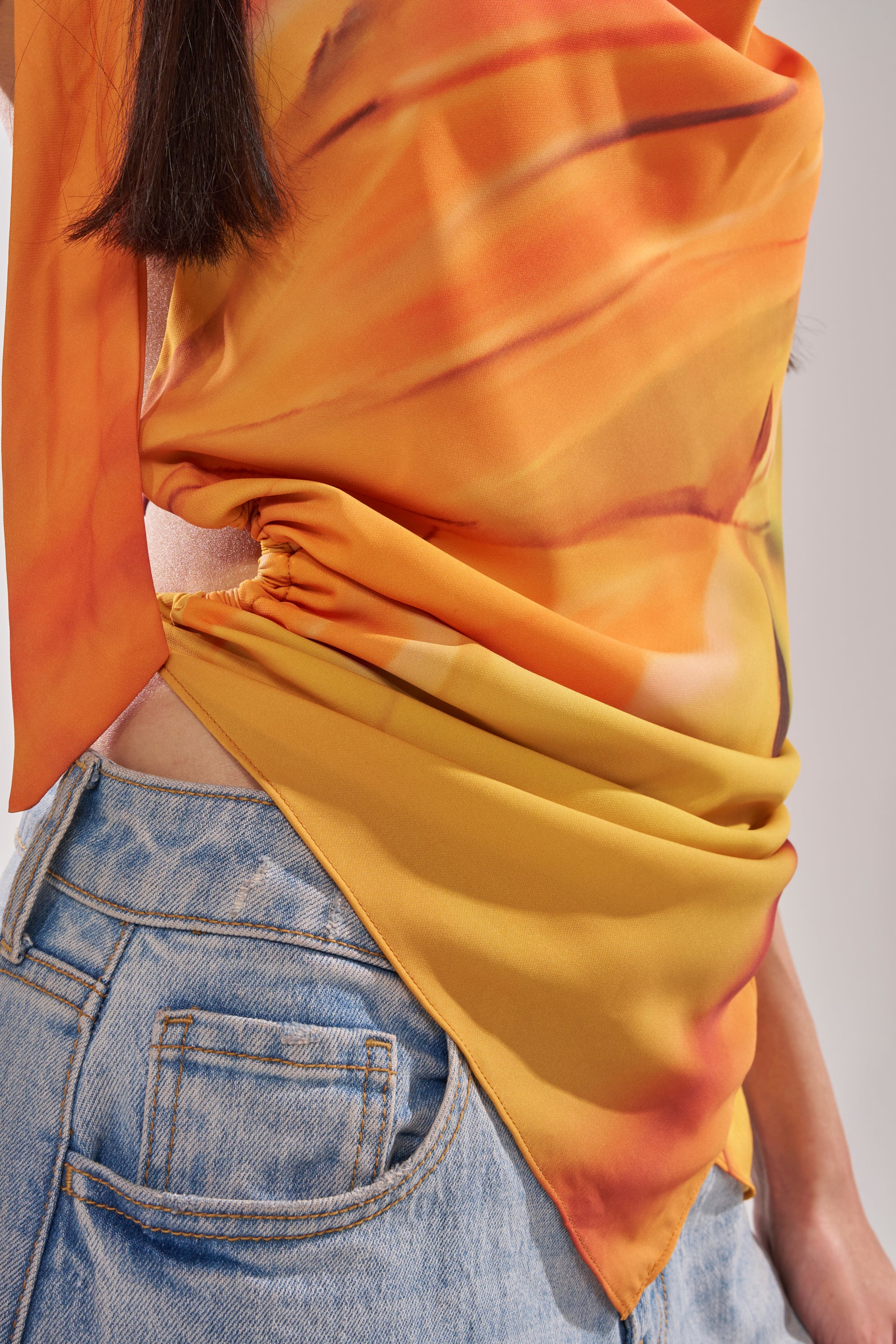 A person wearing the COASTAL HOODED HALTER TOP with a vibrant sunset print and gathered fabric, paired with light blue denim jeans. Only the torso and part of one arm are visible. 12