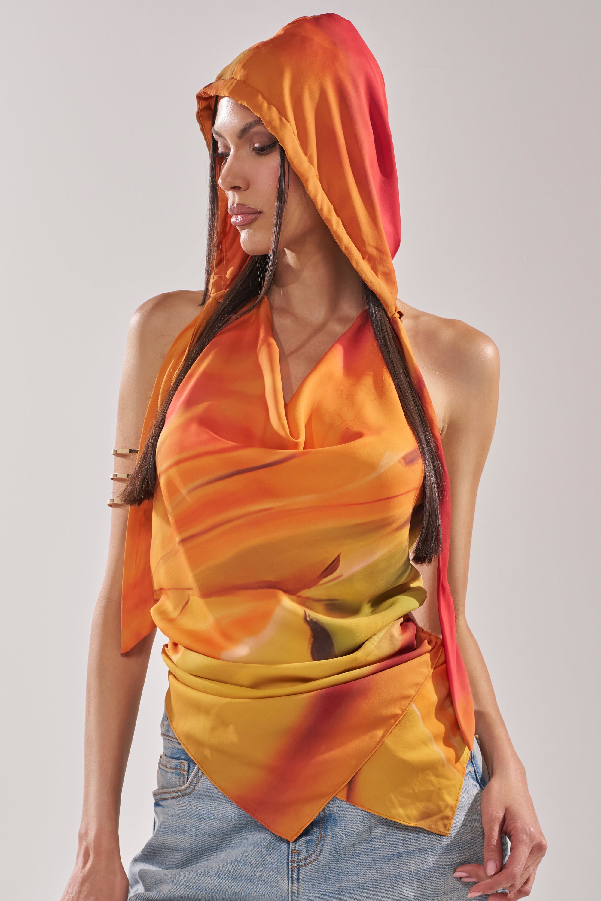 A woman in the COASTAL HOODED HALTER TOP—featuring a vibrant orange and yellow Y2K draped front and hood—pairs it with blue jeans, looking to the side with straight, dark hair. 3