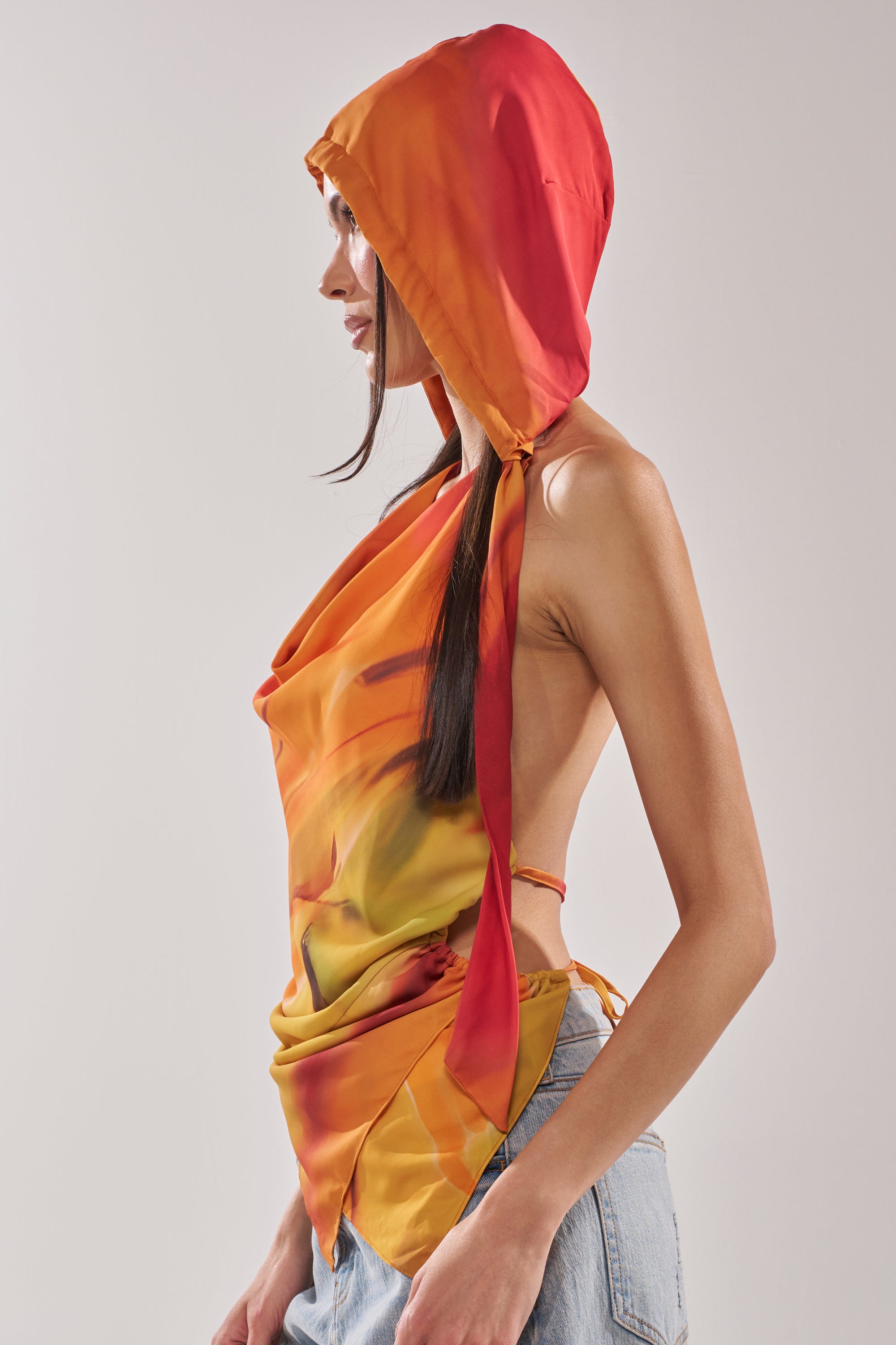 Someone models the COASTAL HOODED HALTER TOP, a vibrant orange and red satin halter with a cowl neckline and hood, paired with light blue jeans, turned sideways against a plain light background. 6