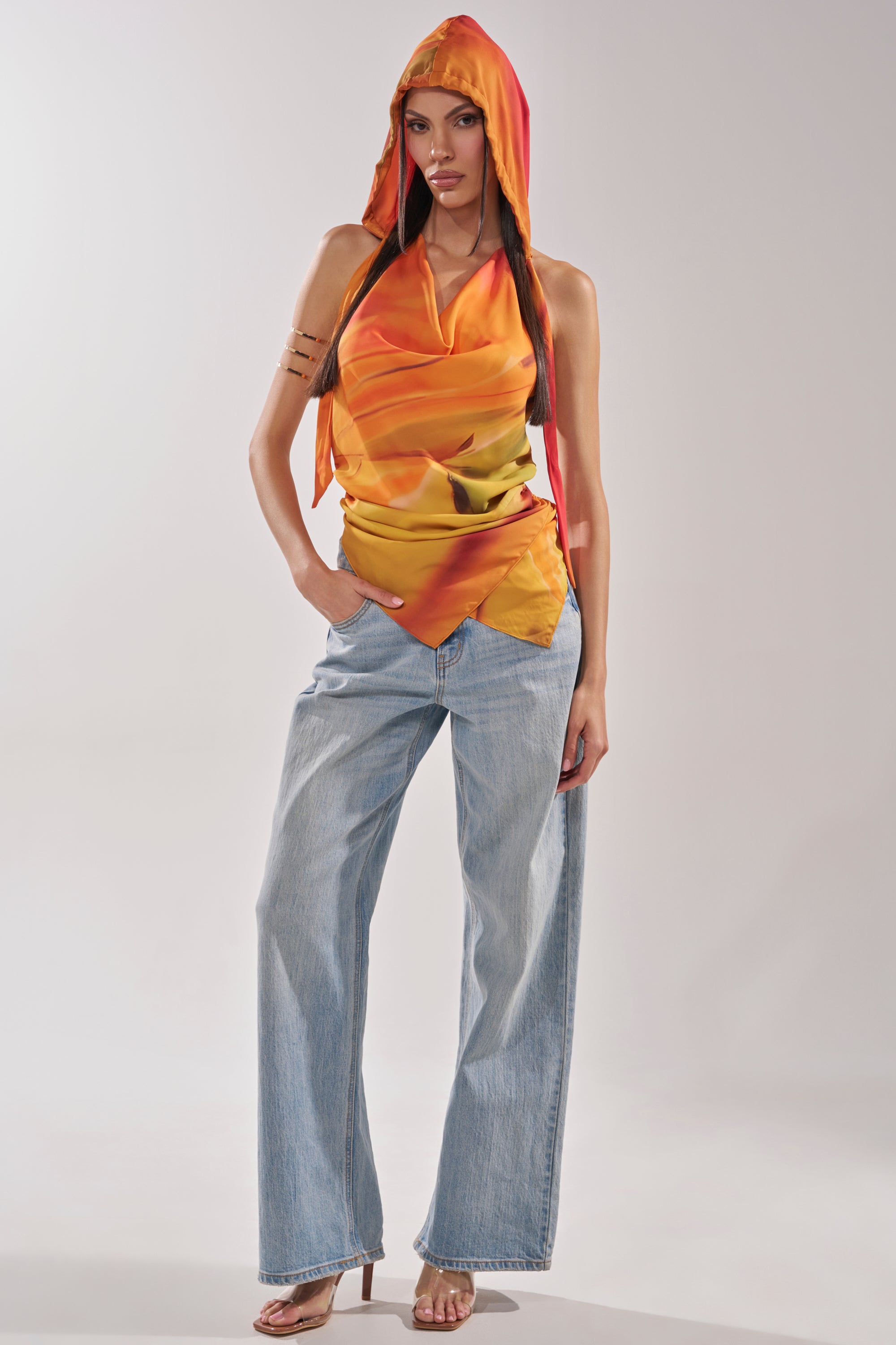 A woman models the COASTAL HOODED HALTER TOP with orange and yellow draped fabric, styled with light blue wide-leg jeans and beige heeled sandals against a plain light background. 8