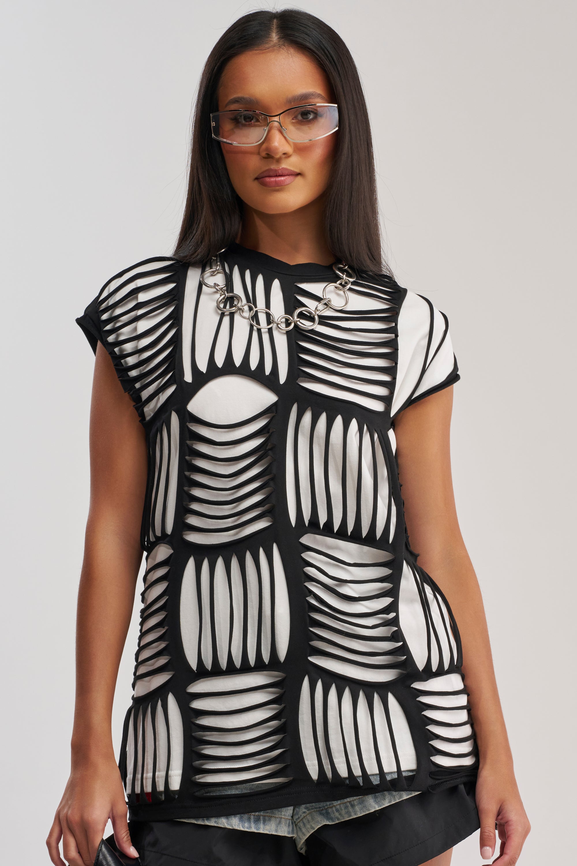 A woman wears large glasses, a chunky chain necklace, and the STRESSED OUT DISTRESSED TEE with a striking black-and-white geometric pattern over black shorts, standing against a plain light background.
