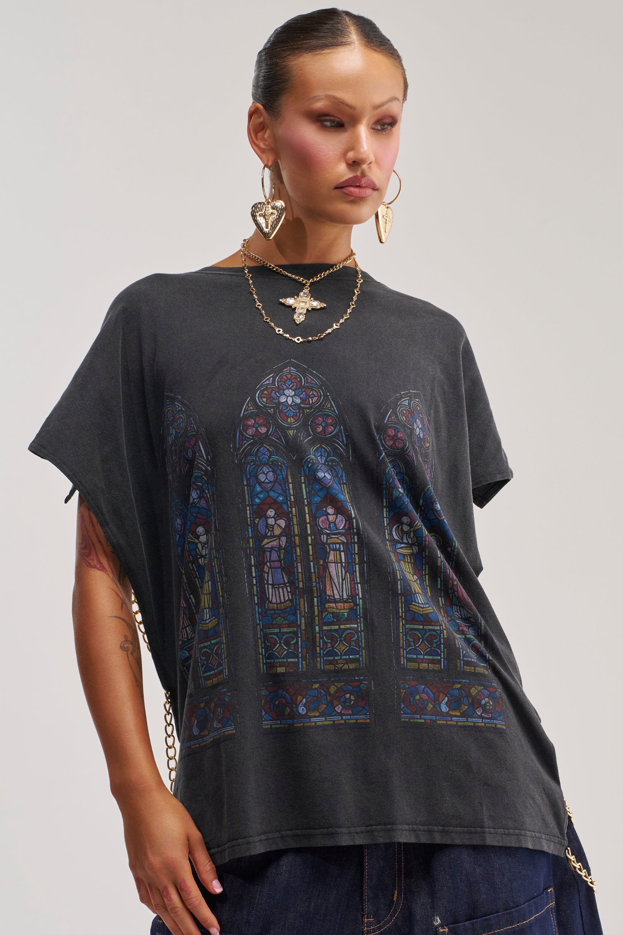 A woman wearing the BLESSED SLEEVELESS MUSCLE TANK in charcoal grey, featuring a stained glass window graphic, styled with a gold cross necklace and heart-shaped earrings. Her slicked-back hair and neutral background complete an effortlessly cool streetwear festival look.