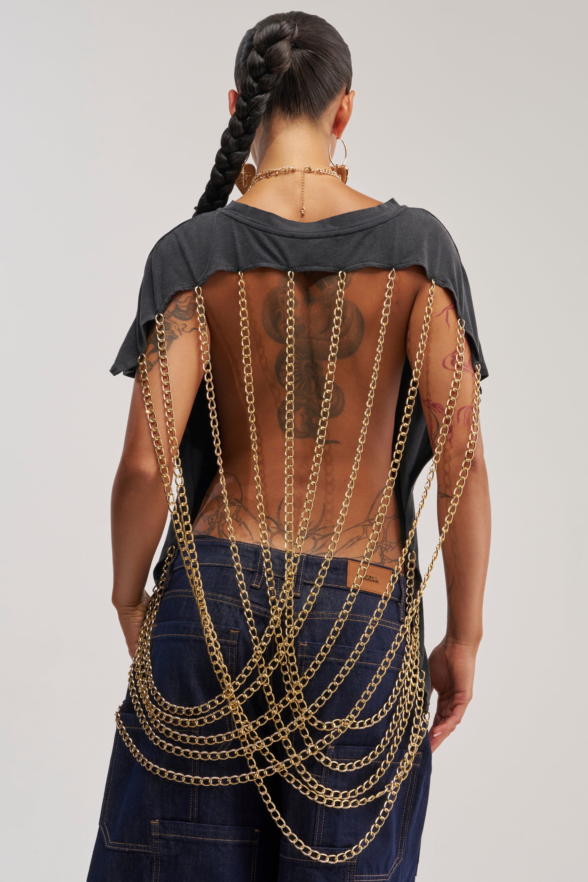 A person with a long braid wears the BLESSED SLEEVELESS MUSCLE TANK styled for a cool streetwear festival vibe, pairing it with blue jeans. Their open back reveals visible tattoos.