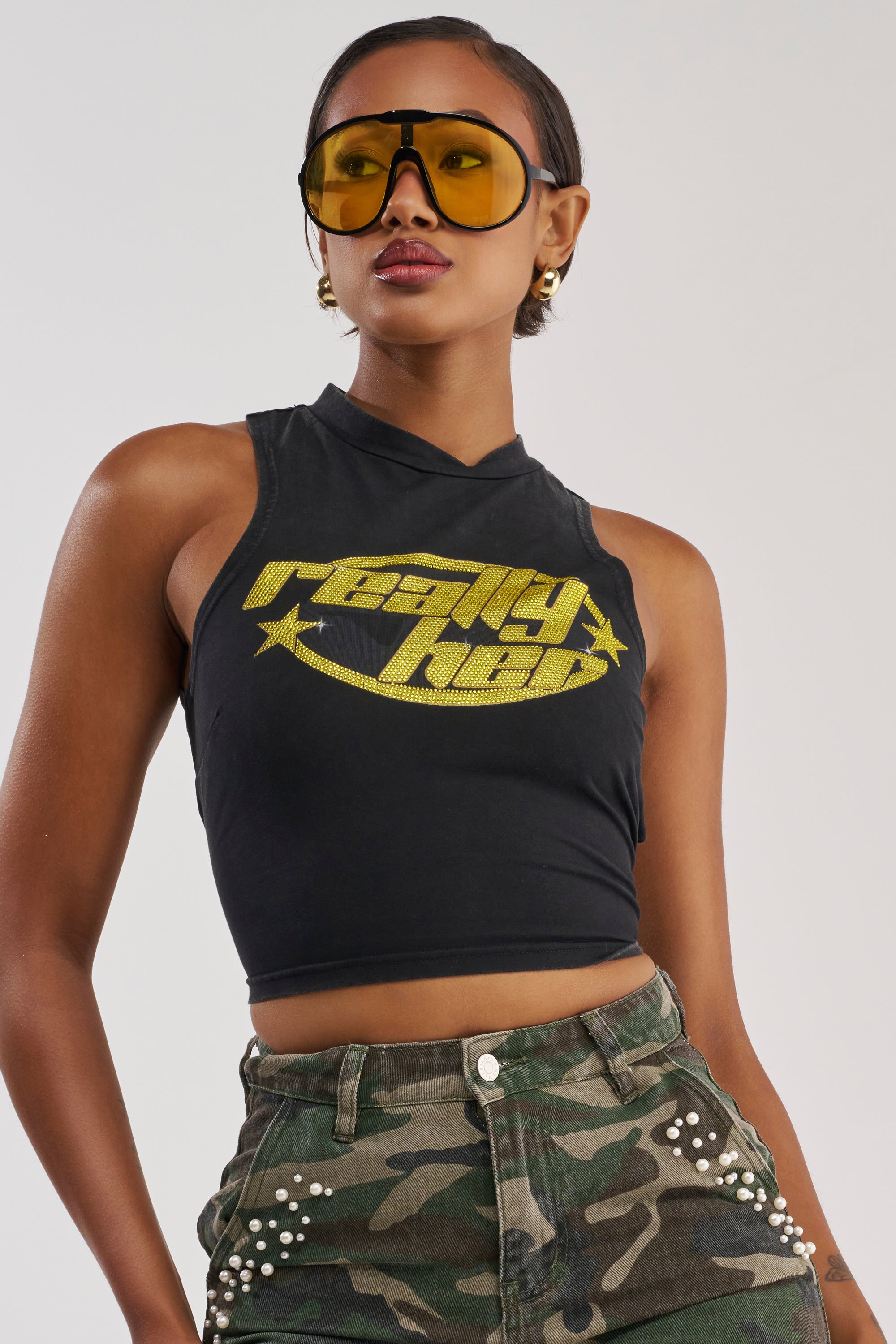 Wearing the REALLY HER TIE-BACK TANK TOP—a sleeveless black crop top with yellow text and stars—paired with large yellow sunglasses and high-waisted camo pants adorned with silver studs, this woman's confident look is the perfect festival outfit against a plain background.