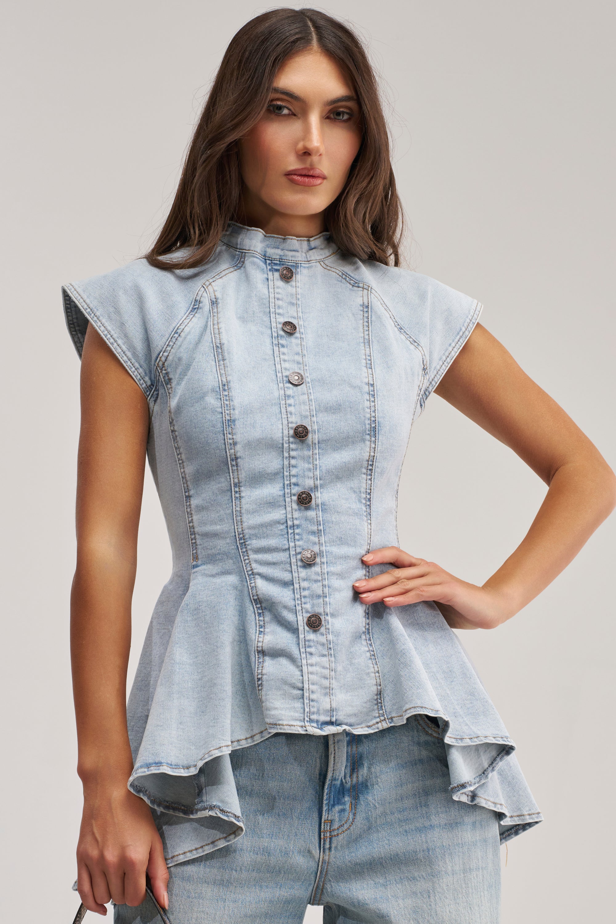 A woman poses in the LUCY DENIM BLOUSE—a sleeveless, light blue button-down with a high collar and flared peplum hem—paired with light washed jeans for easy summer style.