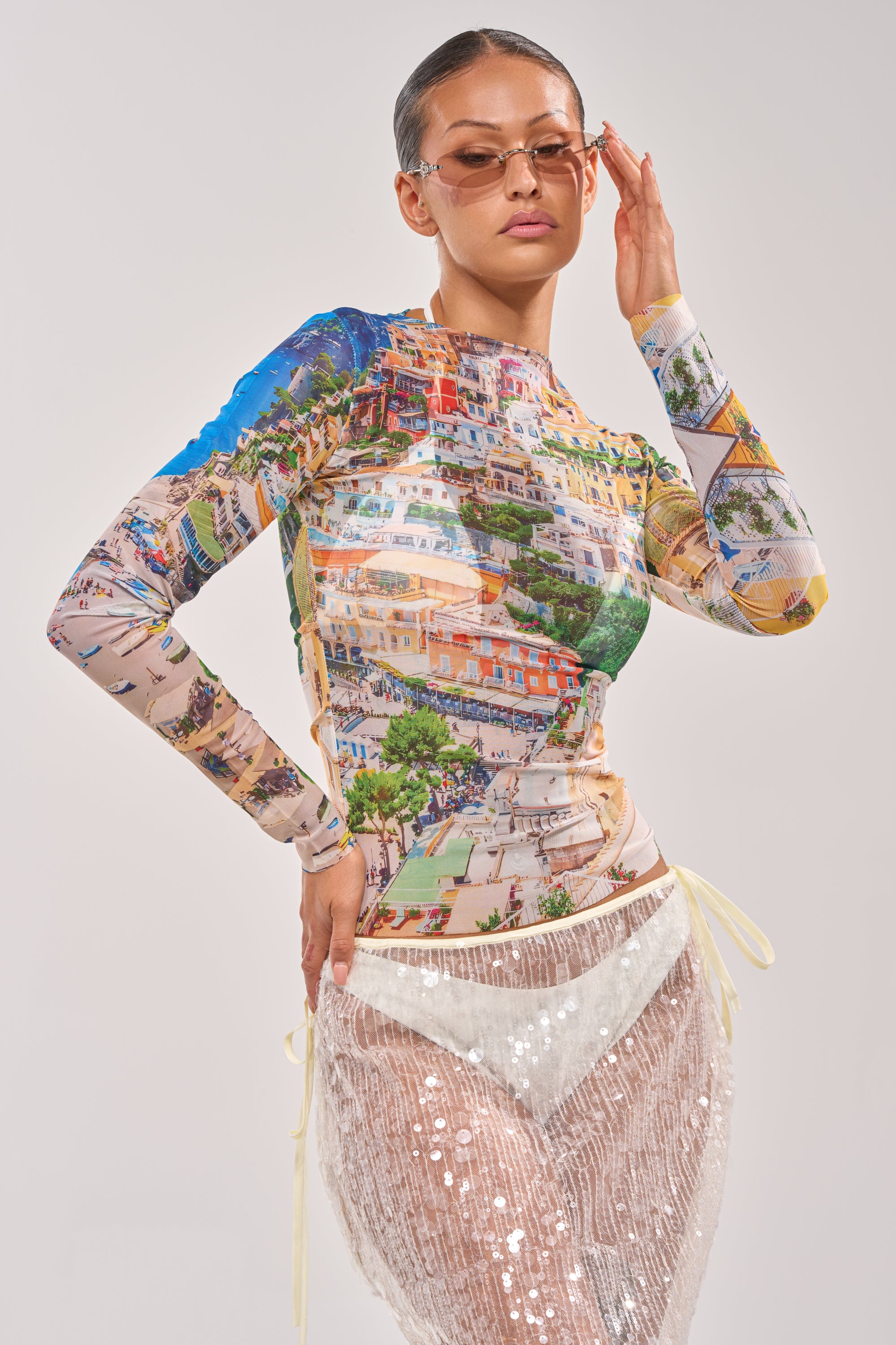 A woman in tinted sunglasses and the AMALFI DAYS MESH TOP, a colorful streetwear-inspired mesh piece with an Amalfi cityscape print, pairs it with a sheer white sequin skirt while posing confidently against a light background. 1