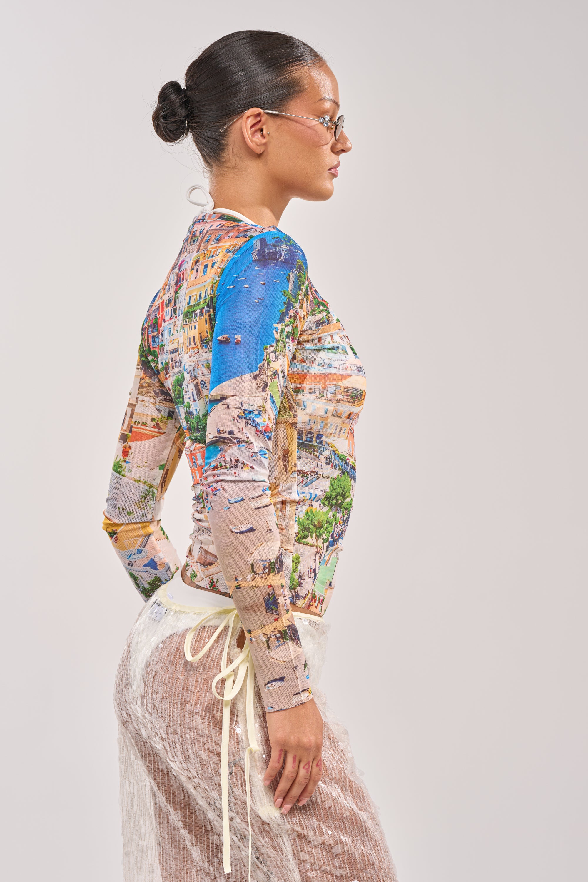 Wearing the AMALFI DAYS MESH TOP, a person with a neat bun and glasses stands in profile against a plain light background, pairing the colorful cityscape mesh top with a sheer white textured skirt. 3