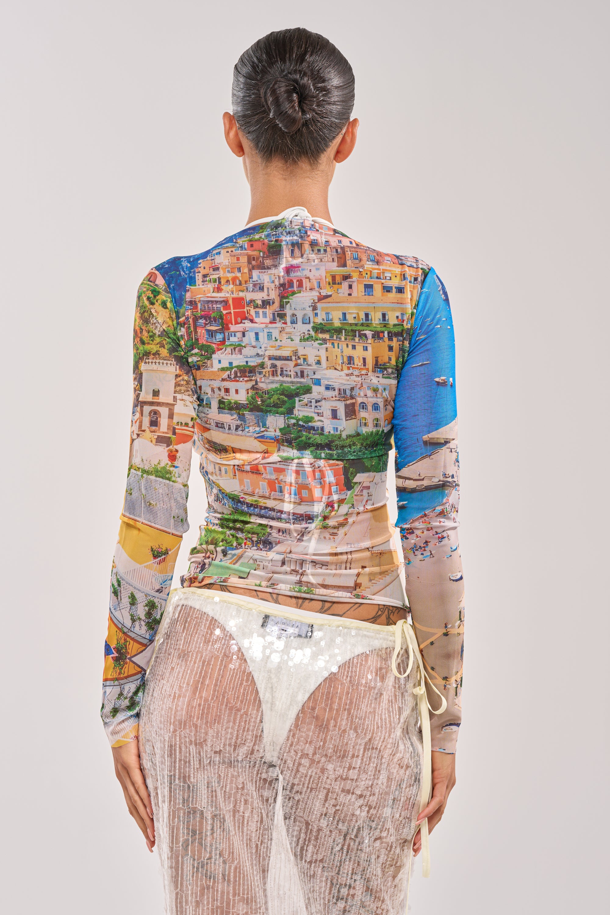 A woman stands with her back to the camera, wearing the AMALFI DAYS MESH TOP featuring a cityscape print and a sheer skirt over white briefs. Her hair is tied in a bun, set against a simple light background. 5