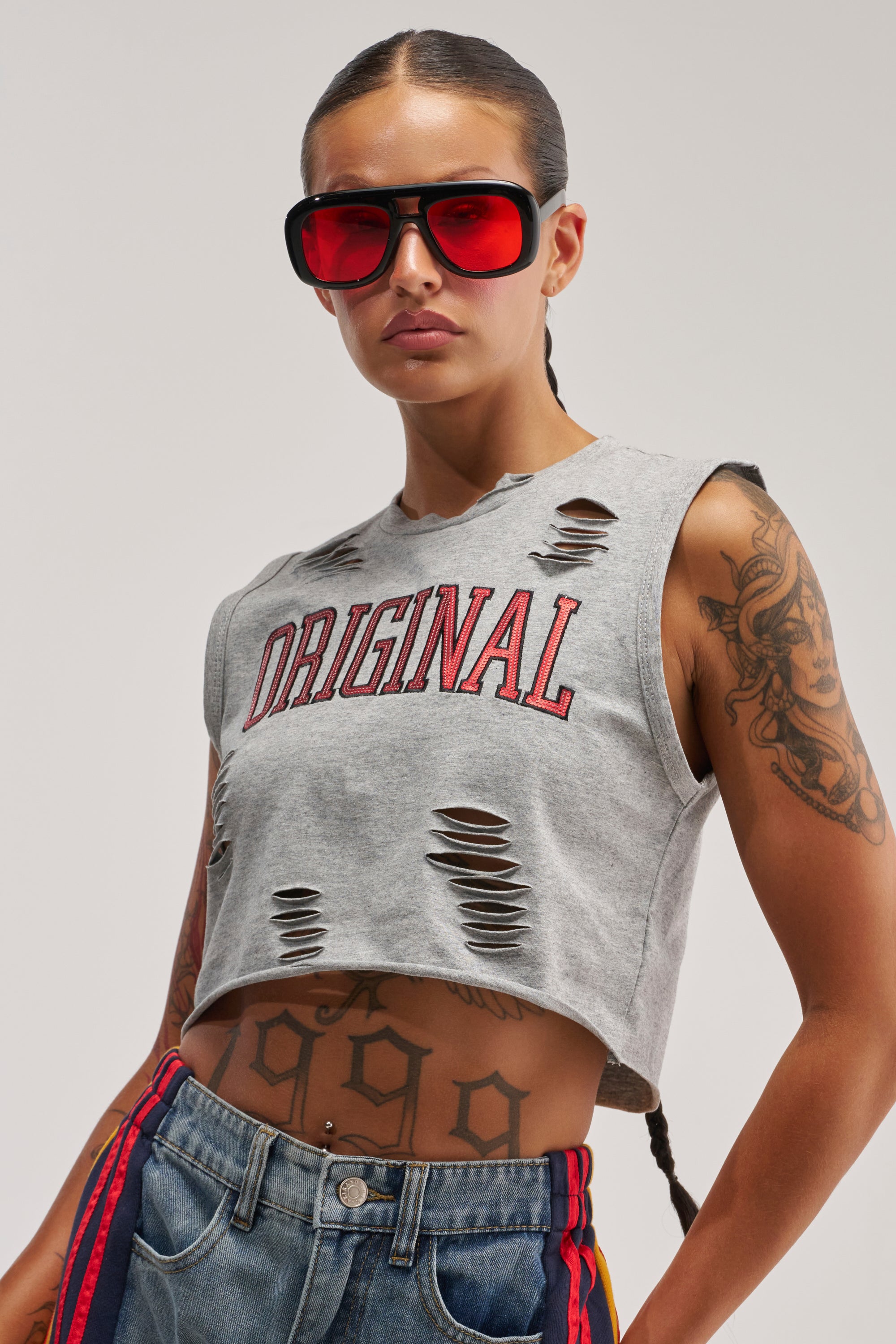 A person in large red sunglasses wears the ORIGINAL SEQUIN CROPPED MUSCLE TEE, a grey top with distressed details and “ORIGINAL” in bold red lettering. Their tattoos, including a prominent “999” on their stomach, are visible beneath the crop. With a long braid and blue jeans featuring striking red and yellow stripes, they complete their streetwear look.