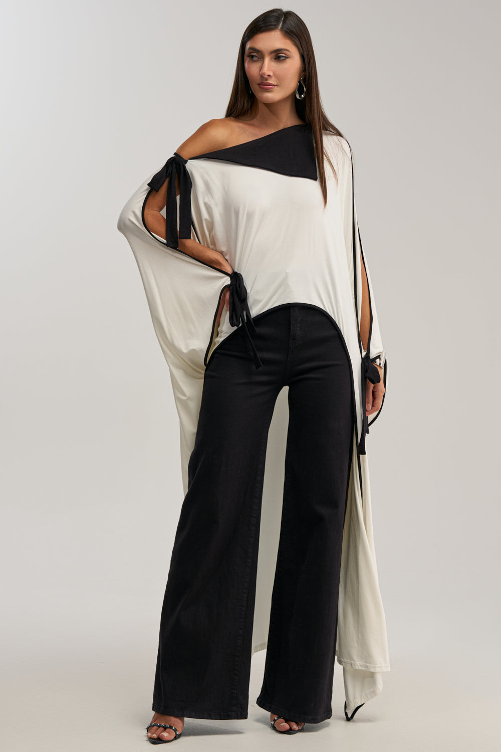 A woman exudes confidence in the EMMA OFF THE SHOULDER CAPE with tied sleeve details, styled with wide-leg black pants and heeled sandals—a chic resort-ready look. She wears her straight brown hair down and opts for minimal jewelry.