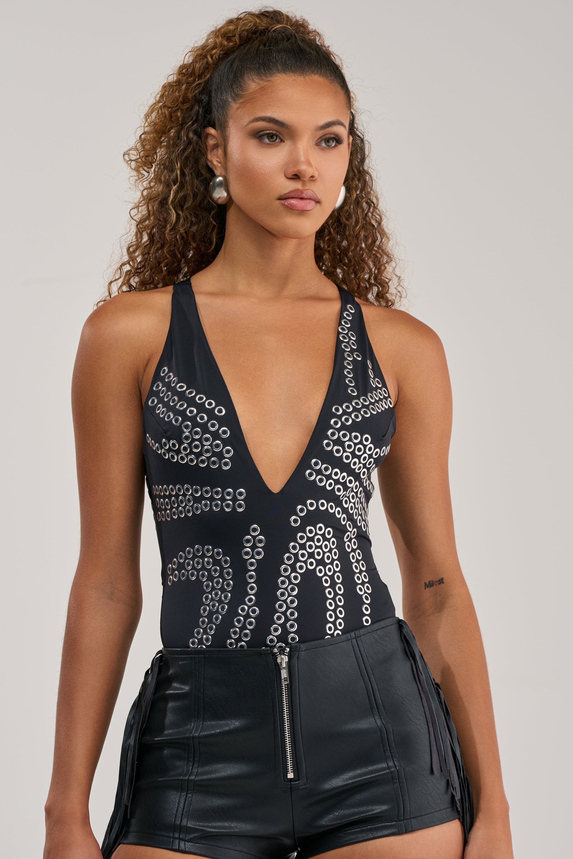 A woman with curly hair models the JENNA GROMMET BODYSUIT—black with silver circular details—paired with high-waisted shorts, creating a bold, standout summer festival look against a plain backdrop.