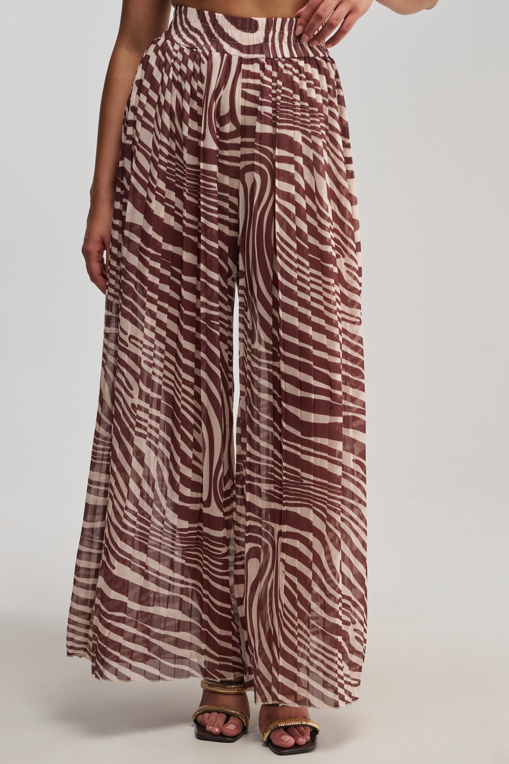 A person wearing the DREAMING OF A VACA PALAZZO PANT, featuring a high-waisted, wide-leg silhouette with brown and cream abstract stripes, pleated fabric, and paired with black sandals—ideal for vacation styling. Only the lower half of the body is visible.