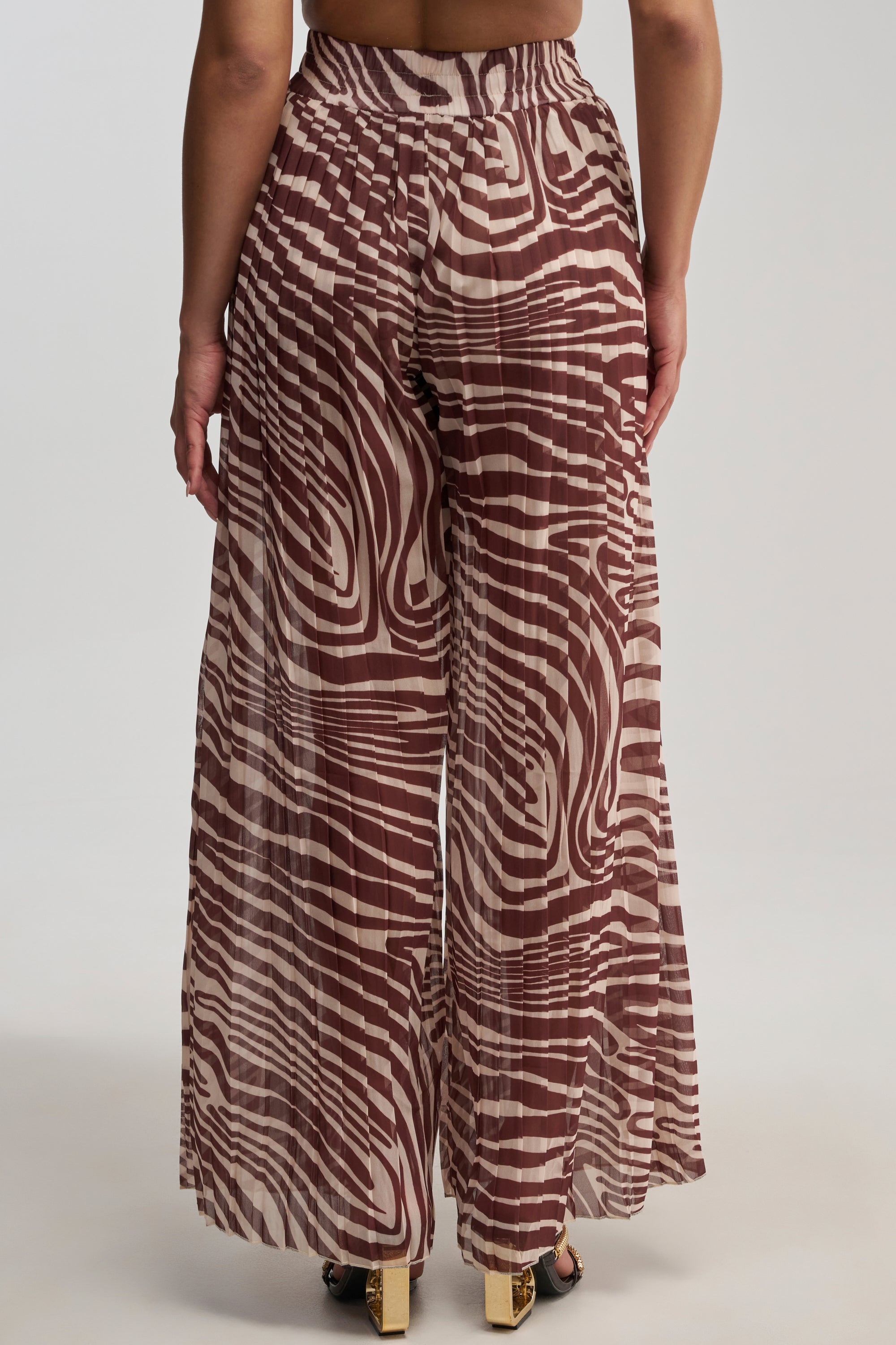 Wearing the DREAMING OF A VACA PALAZZO PANT—featuring wide-leg, high-waisted brown styling with a beige wavy striped pattern—a person is seen from the back against a neutral background, offering perfect inspiration for effortless vacation styling.