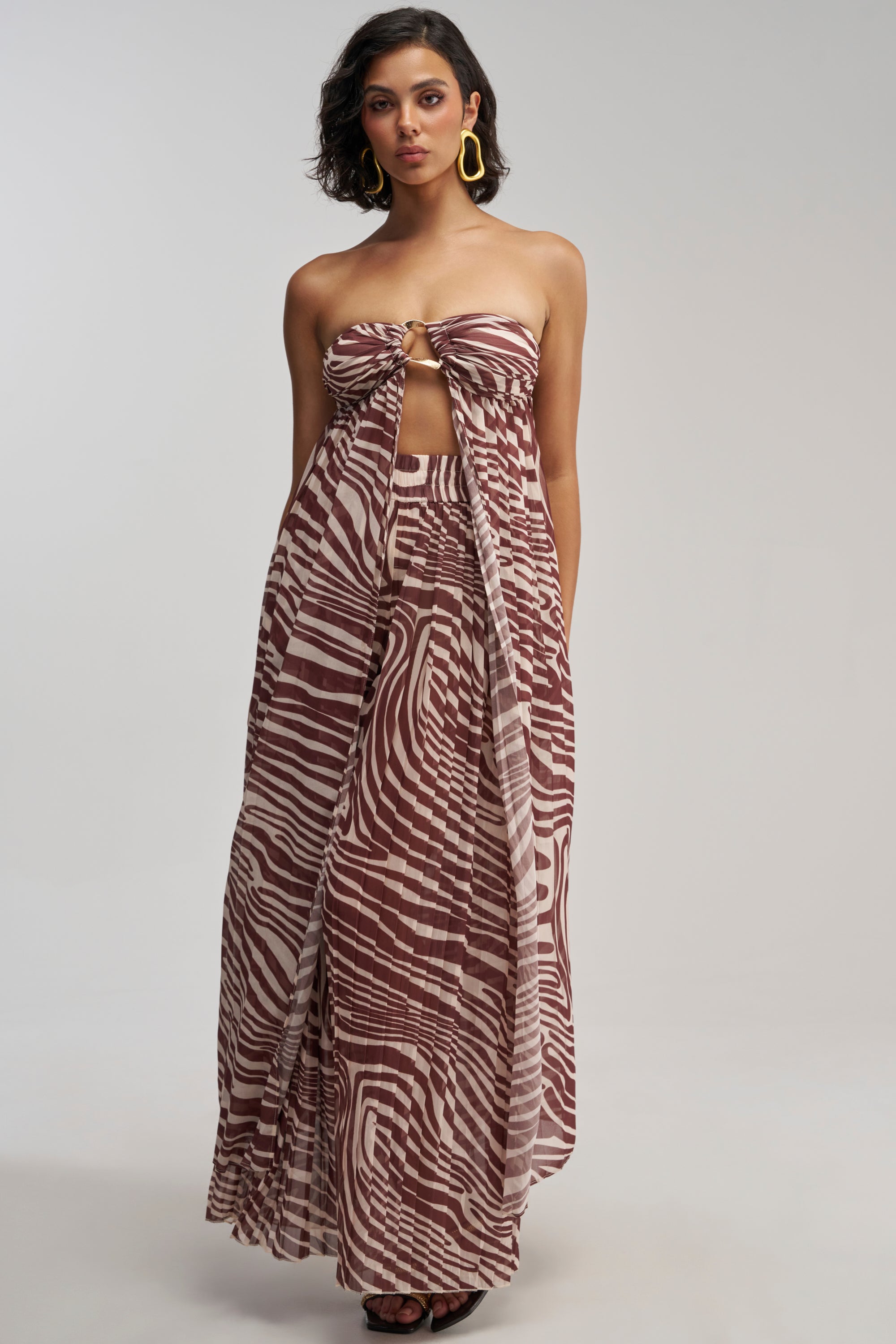 A woman styled in the DREAMING OF A VACA BLOUSE, layered beneath a strapless brown and white patterned maxi dress with a front knot and cutout, accessorized with large yellow earrings, poses against a plain light background.