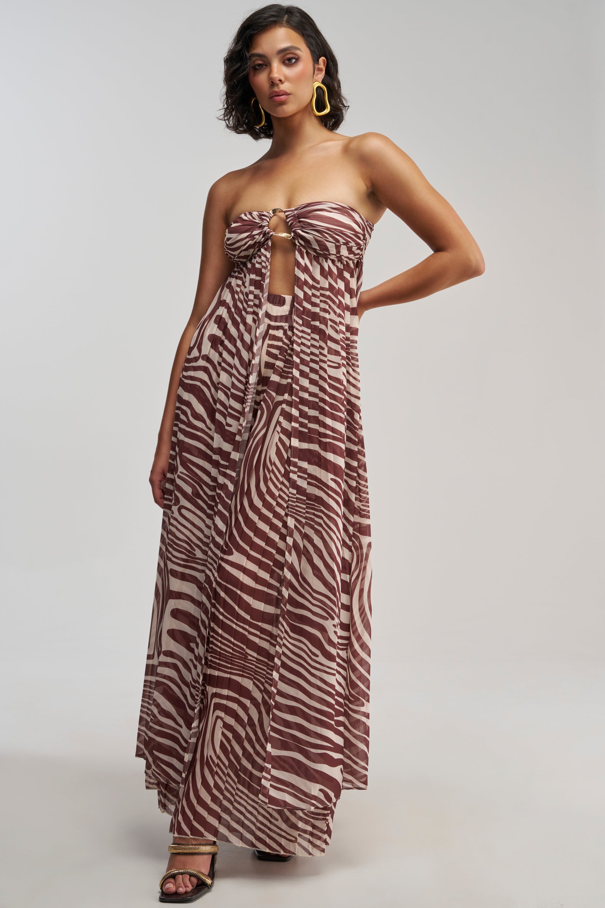 A woman wears the DREAMING OF A VACA BLOUSE, a strapless brown and white zebra-print longline blouse featuring a front knot and cutout, styled as a maxi dress. She pairs it with yellow hoop earrings and black heeled sandals, standing against a plain background.