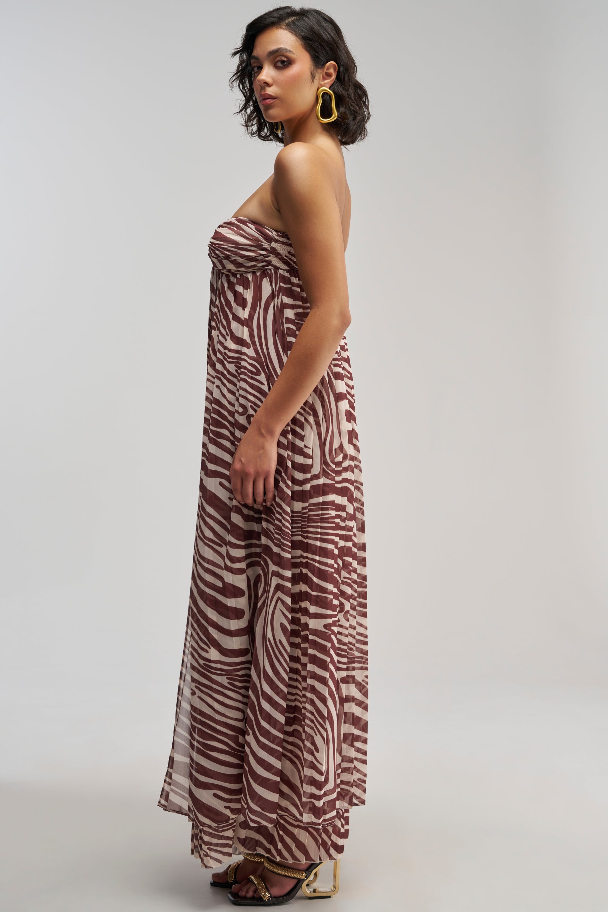 Woman in the DREAMING OF A VACA BLOUSE—a bold, strapless, floor-length top with brown and white zebra stripes—styled with yellow hoop earrings and high-heeled sandals as she poses sideways against a plain backdrop, exuding vacation-ready flair.