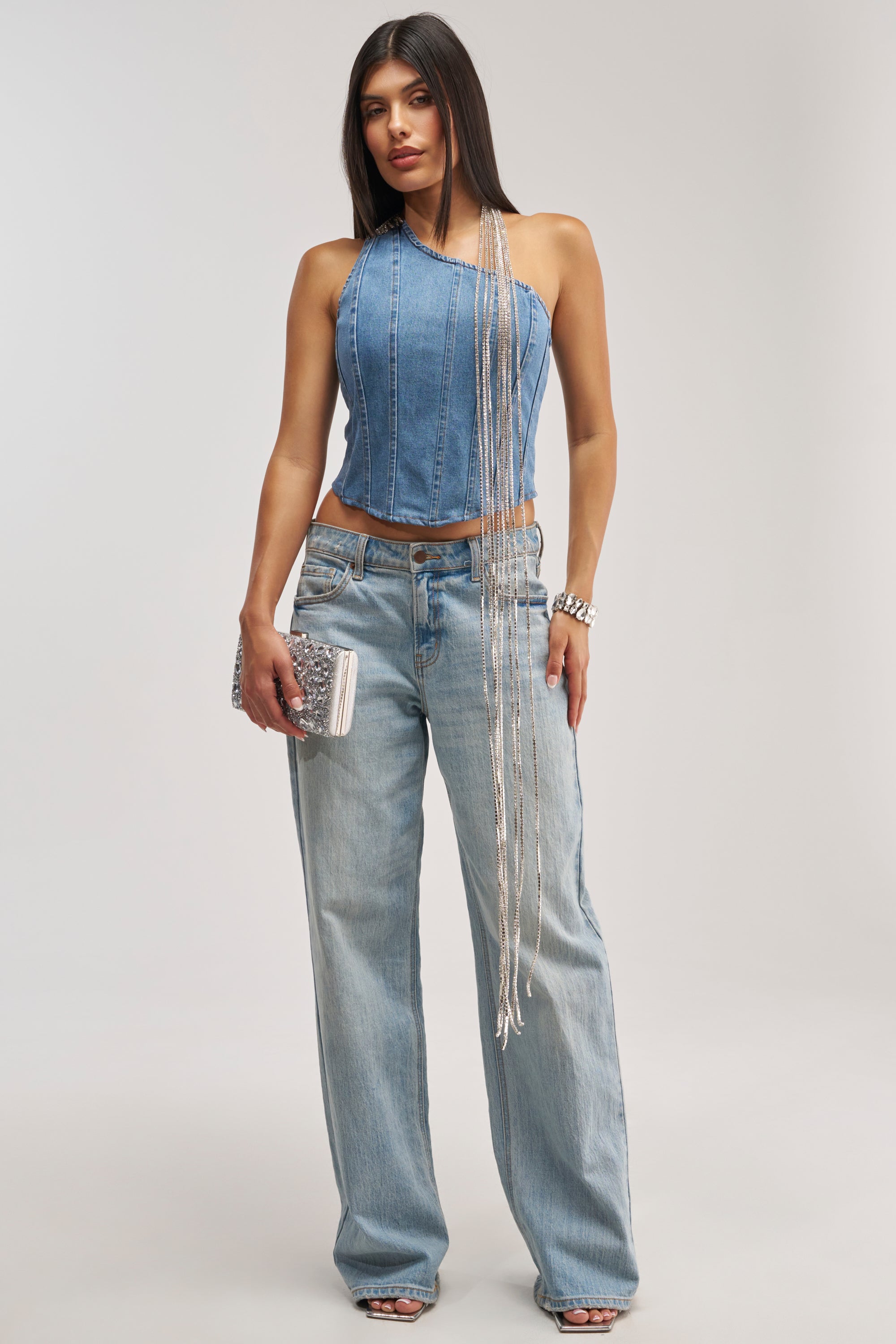A woman wears the YOU WOULDNT GET IT RHINESTONE DETAIL CORSET, styled with light blue jeans and a silver clutch, accessorized with layered necklaces and bracelets, standing against a plain light background. This chic ensemble makes the perfect festival outfit with its rhinestone accents and effortless style.