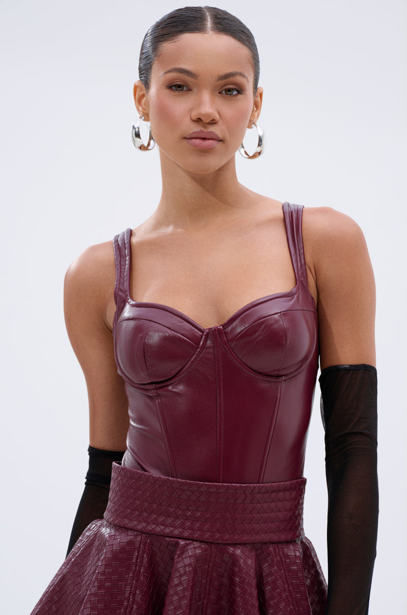 A woman with slicked-back hair wears the EASY LOVER BODYSUIT IN PLUM, a matching textured skirt, large silver hoop earrings, and sheer black gloves, standing against a plain light background. 0