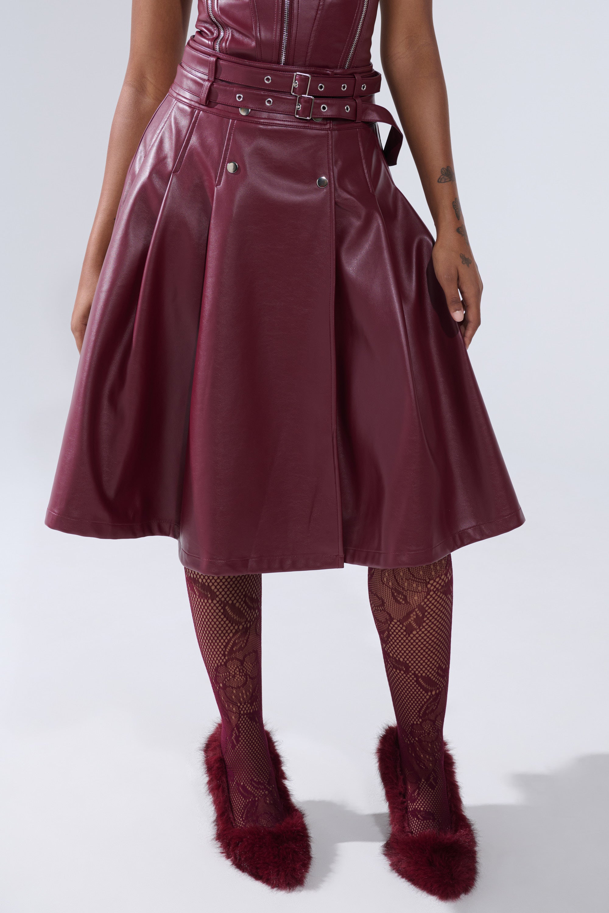 A person stands against a plain light background, wearing the FEELING THE ENERGY MIDI SKIRT in shiny burgundy faux leather with a matching belt, paired with patterned burgundy tights and fuzzy slippers. Image cropped from shoulders down.