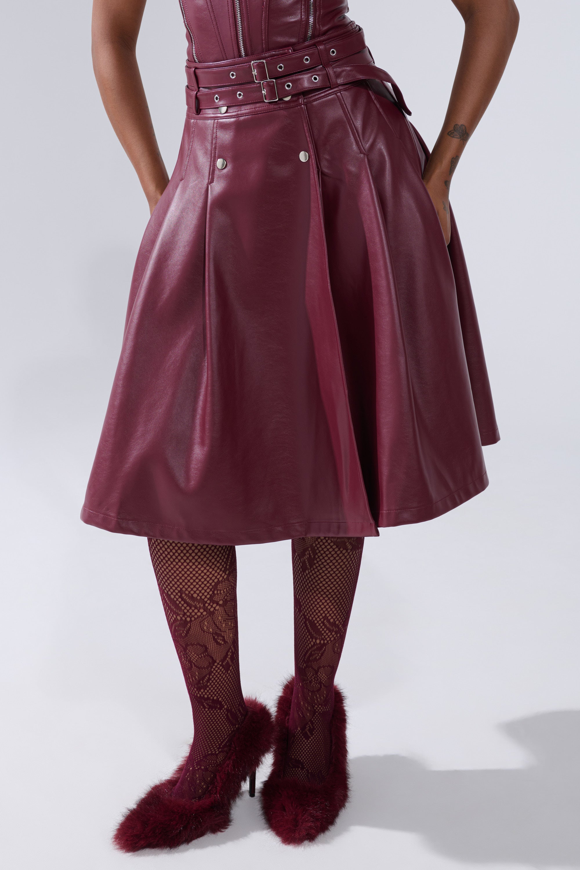 Wearing the FEELING THE ENERGY MIDI SKIRT in burgundy faux leather with a belted waist, lace-patterned tights, and matching fuzzy high-heeled shoes, the person stands with hands in pockets; upper body partially out of frame.